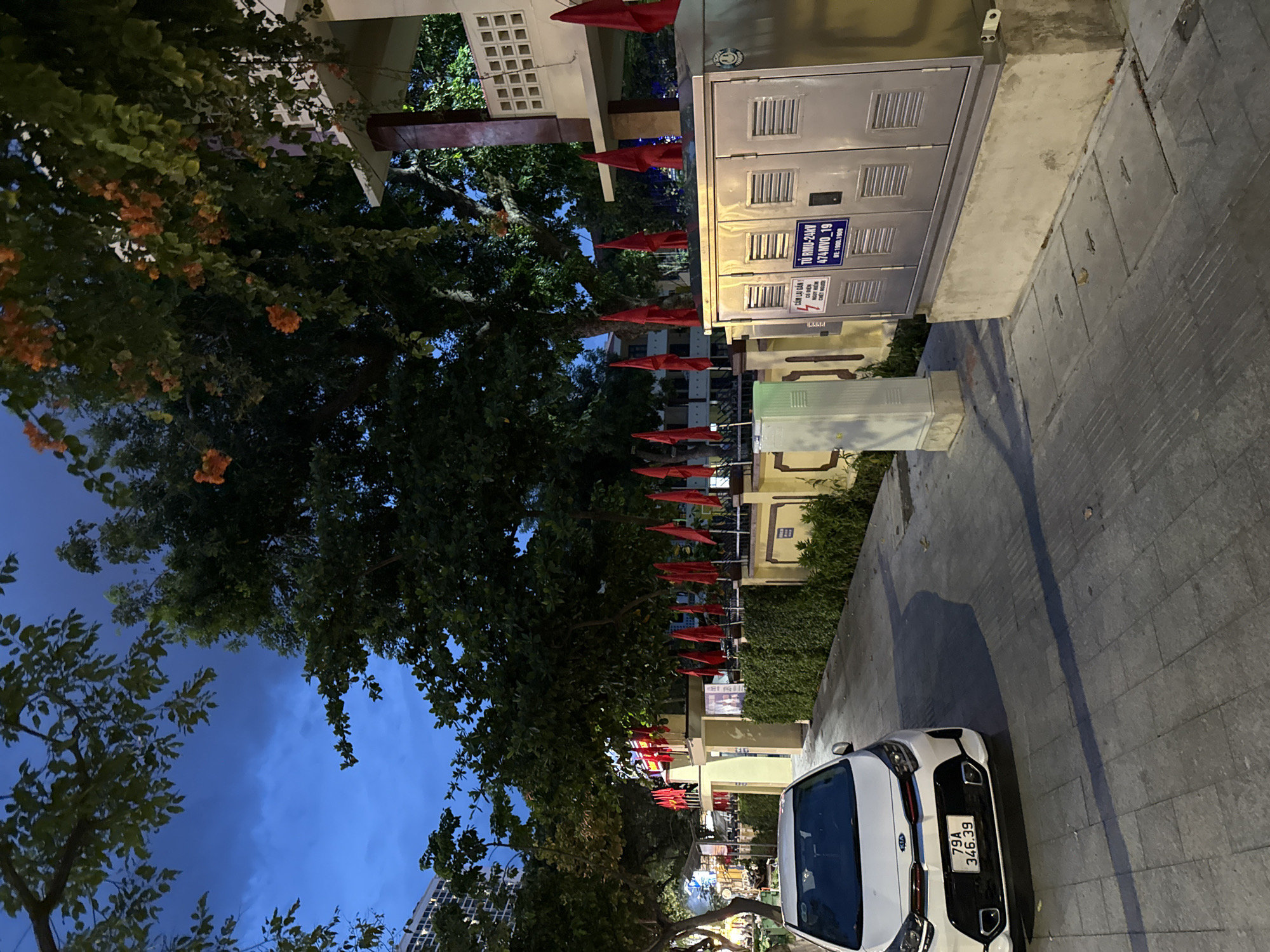 This is a daytime photograph taken outdoors, depicting a street scene in Nha Trang, Vietnam. The foreground features the front half of a white Kia car parked on a paved sidewalk. The car has a license plate that reads "79A 346.39". Adjacent to the sidewalk is a metal electrical utility box with labels and vents. Further back, a row of red flags are displayed along a low wall and interspersed with trees. Behind the flags, a building with white latticework on its exterior is visible, along with dense green foliage. The sky above is a deep blue, suggesting it might be evening or twilight. The overall environment appears to be a well-maintained urban or suburban area, possibly a public space or along a promenade.