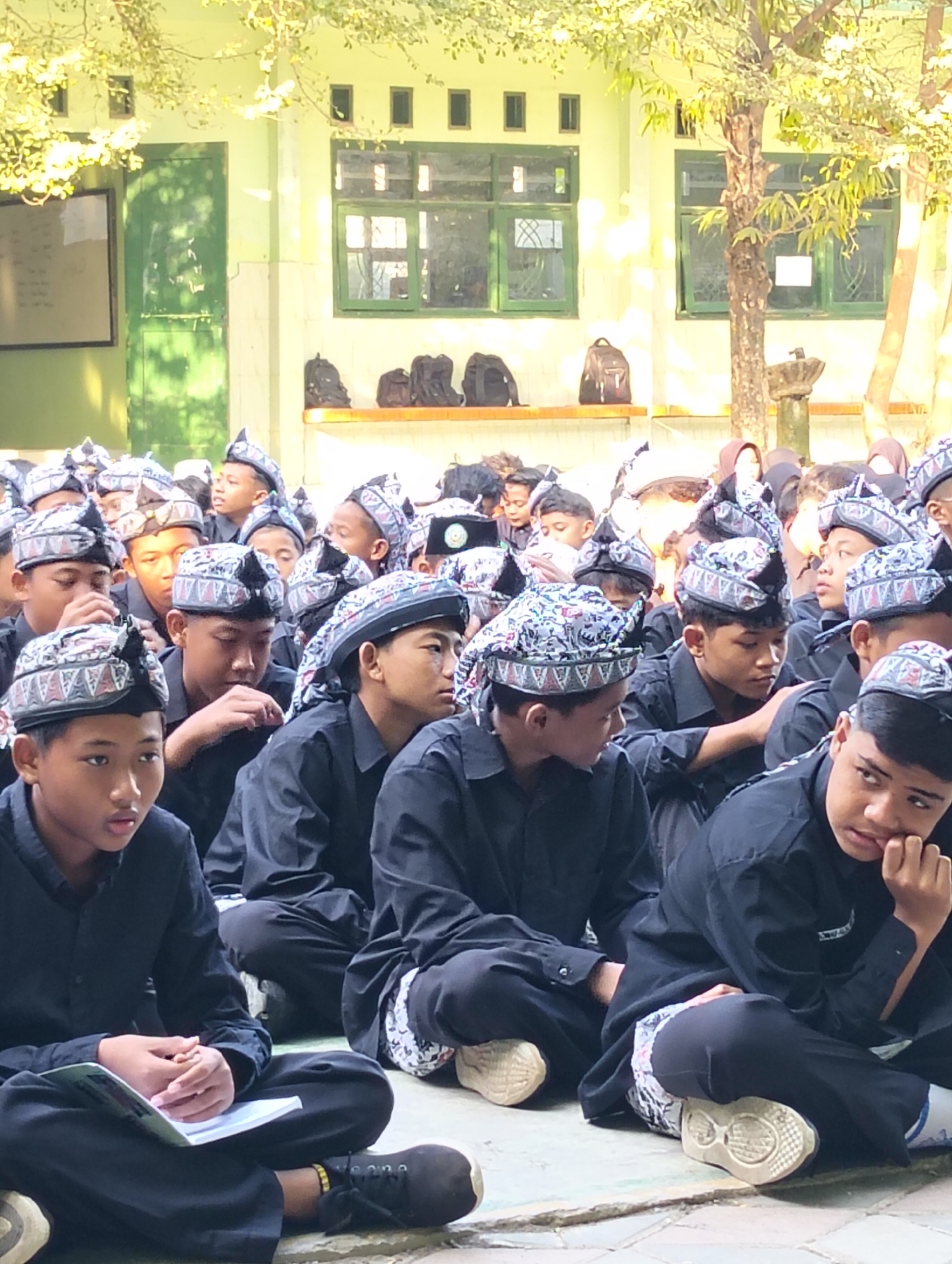A group of young boys, likely students, are seated on the ground in front of a school building. They are all wearing black shirts and pants, and most are adorned with traditional patterned headwear. The building behind them appears to be a classroom with multiple windows. Several backpacks are visible on a ledge along the building. The ground is paved with stone. The scene suggests an outdoor gathering or event at a school in Dagan, Indonesia.