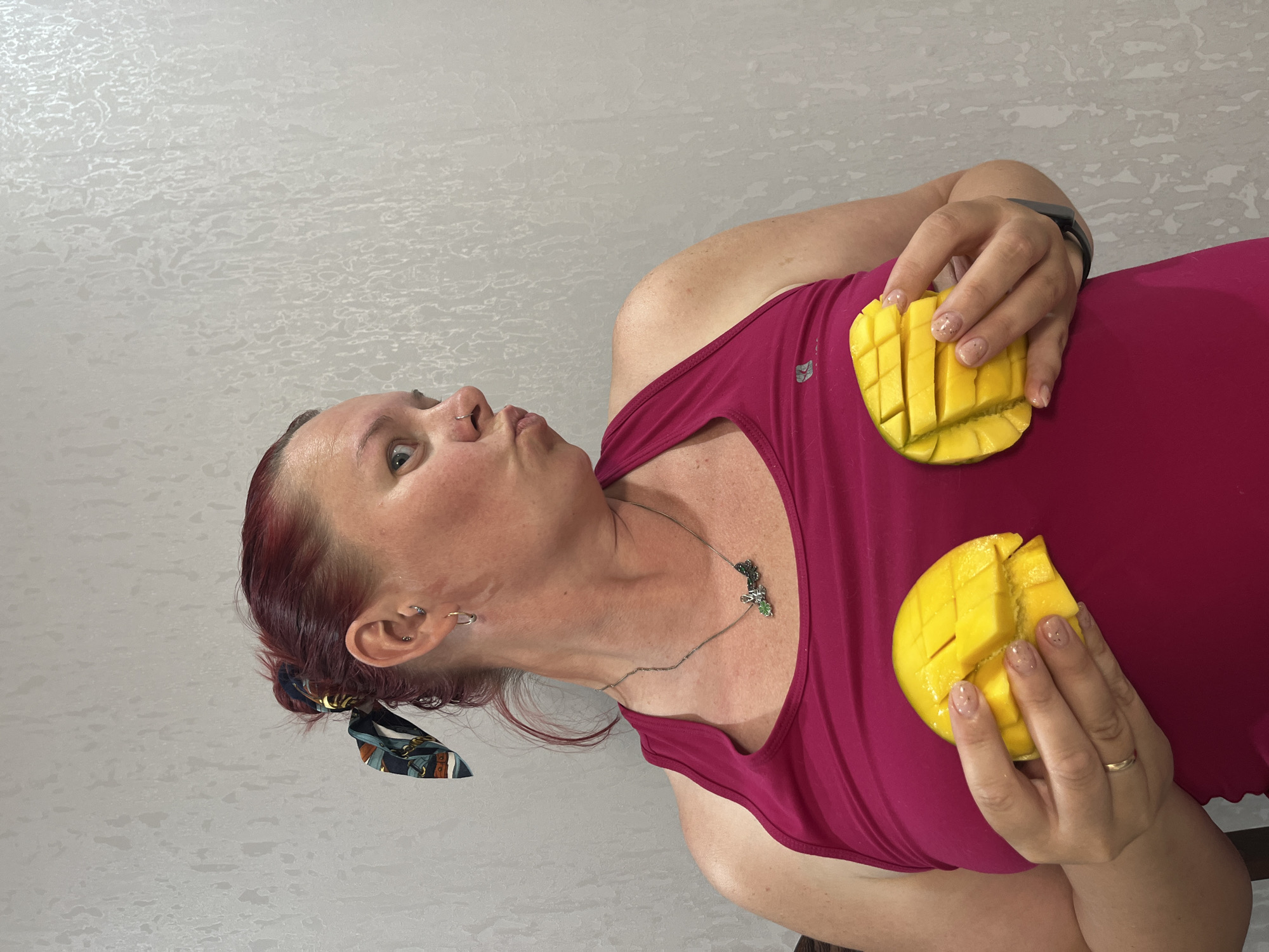 A woman is holding two mangoes cut into grid patterns. She is wearing a red tank top and a necklace with a dragonfly pendant. Her hair is reddish and tied back with a patterned scarf. She is indoors and the background is a light-colored textured wall. It is not possible to confirm whether the image was taken in Chaikovsky, Russia.