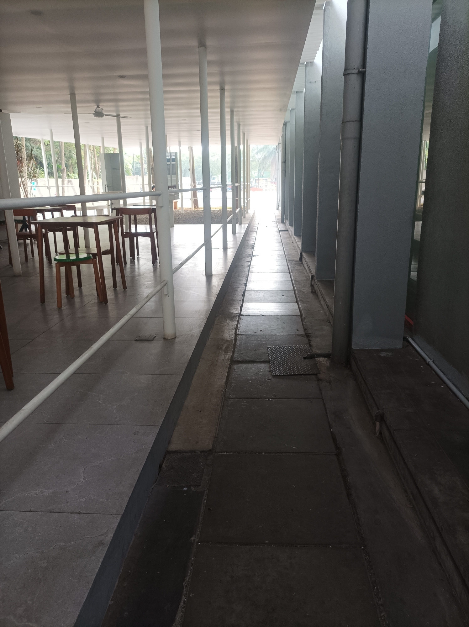 The image depicts an outdoor covered walkway in South Jakarta, Indonesia. The walkway features a tiled floor with a distinct change in material and elevation at the halfway point. On the left side, there are wooden tables and chairs arranged under a white roof supported by white cylindrical pillars. A ceiling fan is visible in the distance. The right side of the walkway has a drain channel running alongside grey, rectangular pillars. The scene is brightly lit from the end of the walkway, suggesting daylight. There are no people visible in the image. The environment appears to be an open-air seating or dining area, possibly part of a restaurant or public space.