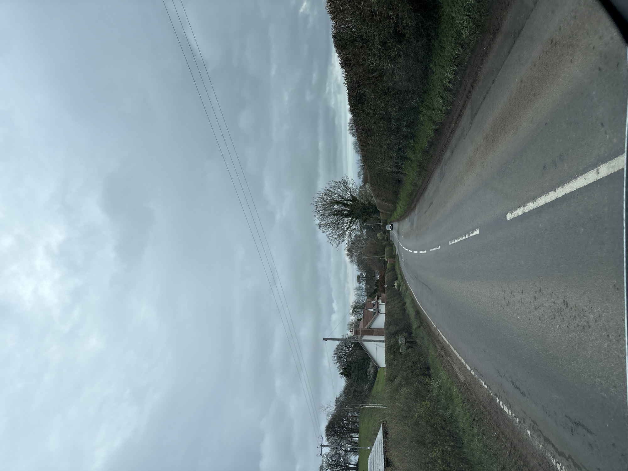 The image depicts a rural asphalt road in Ruyton-XI-Towns, United Kingdom, under an overcast sky. The view is presented from a low perspective, indicative of an occupant inside a vehicle, looking forward. The sky is uniformly covered with light grey clouds.
The road curves gently to the right in the mid-distance. It features a continuous white line marking the left edge and broken white lines indicating the center. The asphalt surface exhibits scattered damp patches. Both sides of the road are bordered by muddy verges, which transition into dense, dark green hedges.
On the left side of the road, behind the hedge line, a white residential building with a brown tiled roof and a prominent brick chimney is visible. Behind this structure, a grassy hill rises, populated with bare deciduous trees and some evergreen foliage. A dark utility pole with at least two power lines extending horizontally across the frame is present on the left. A small, dark green sign with white text, partially obscured by grass, is visible on the left verge.
On the right side of the road, a similar dense, dark green hedge runs parallel to the roadway. Further down the road, behind the hedge, a large, bare deciduous tree with a spreading canopy stands. A dark-colored vehicle is visible in the distance on the road, proceeding away from the viewer. No individuals or direct interactions are discernible within the scene. The environment suggests a temperate, possibly winter or early spring, setting due to the bare trees.