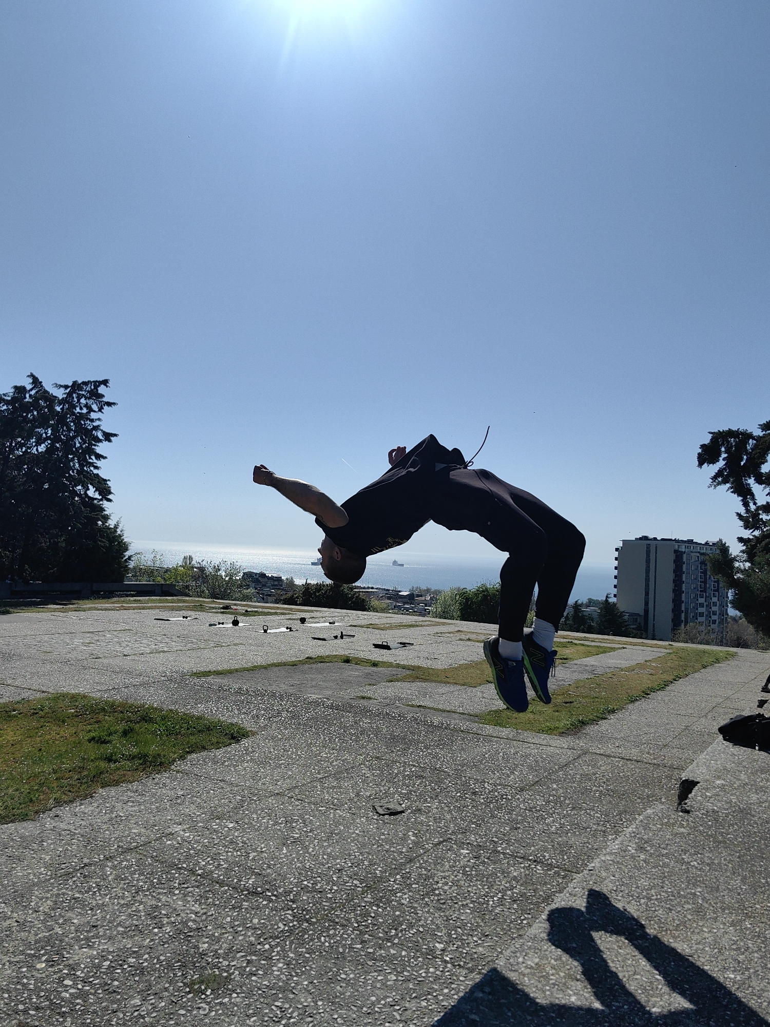 A person is in mid-air performing a backflip. The person is wearing a black shirt, black pants, white socks, and blue and green athletic shoes. The person's arms are extended and the body is arched backwards. The background shows a clear blue sky, a body of water with a ship visible on the horizon, a city with buildings, and trees. The person is on a concrete platform with patches of grass. The photograph was taken outdoors in Unknown.