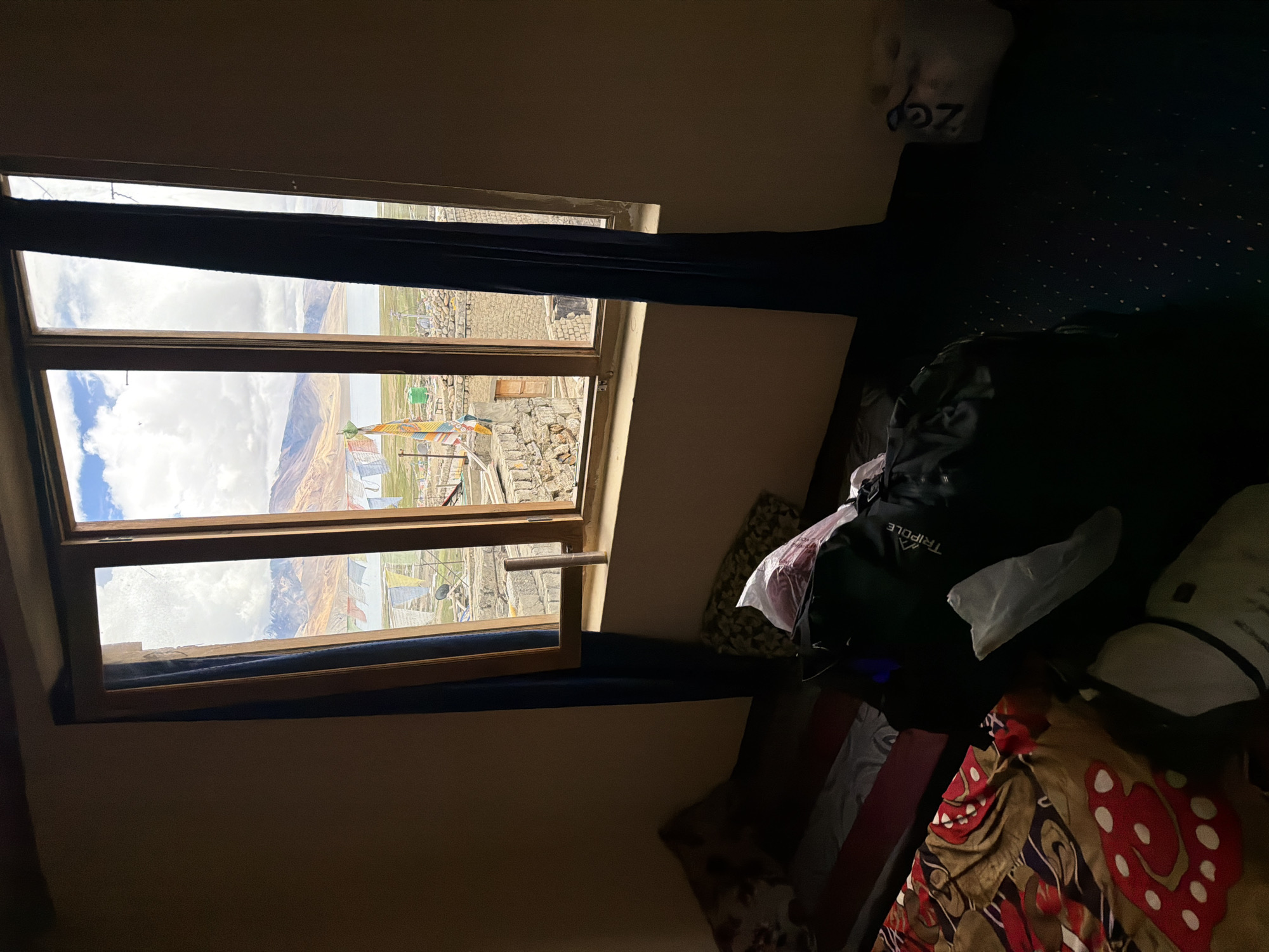 The image captures a view from a room in Karzok, India, looking out a multi-paned window. The window reveals a scenic landscape featuring a body of water, likely a lake, flanked by mountains. Prayer flags, typical of Tibetan Buddhist culture, are strung across the foreground, adding a splash of color to the scene. The room itself appears lived-in, with a bed on the left side adorned with a patterned duvet. A black backpack with the word "TRIPOLE" visible on it is placed on the floor in the lower right quadrant of the image, alongside other bags and a white plastic bag with writing on it. The walls of the room are a neutral tone, and dark blue curtains frame the window. The overall impression is one of a simple, functional living space with a stunning natural backdrop.