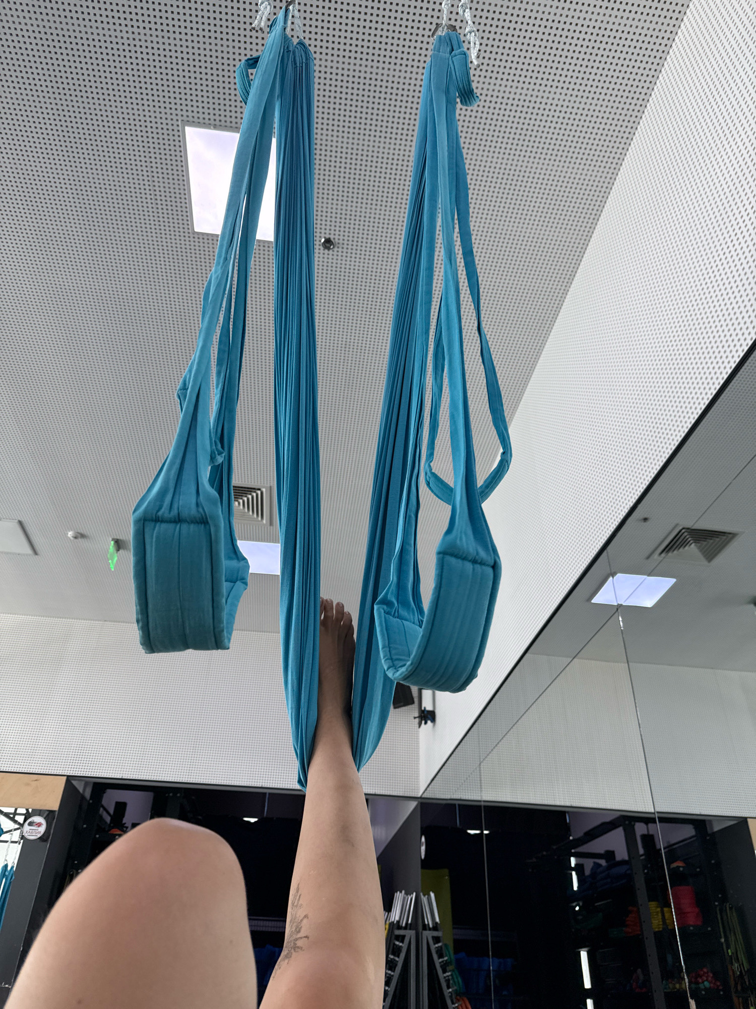 The image shows a person's legs and torso in the foreground, with the person hanging upside down in a teal aerial yoga hammock. The hammock is suspended from the ceiling, which is white and perforated. There are two aerial yoga hammocks visible, one on the left and one on the right. The person's right leg is bent at the knee and resting in the right hammock, while their left leg is extended upwards and out of frame. A portion of the person's thigh and knee is visible in the lower left corner of the image. In the background, a large mirror reflects gym equipment, including weight racks and other exercise machines. The ceiling has square lighting panels, and ventilation grates are visible. The overall environment appears to be an indoor fitness studio.