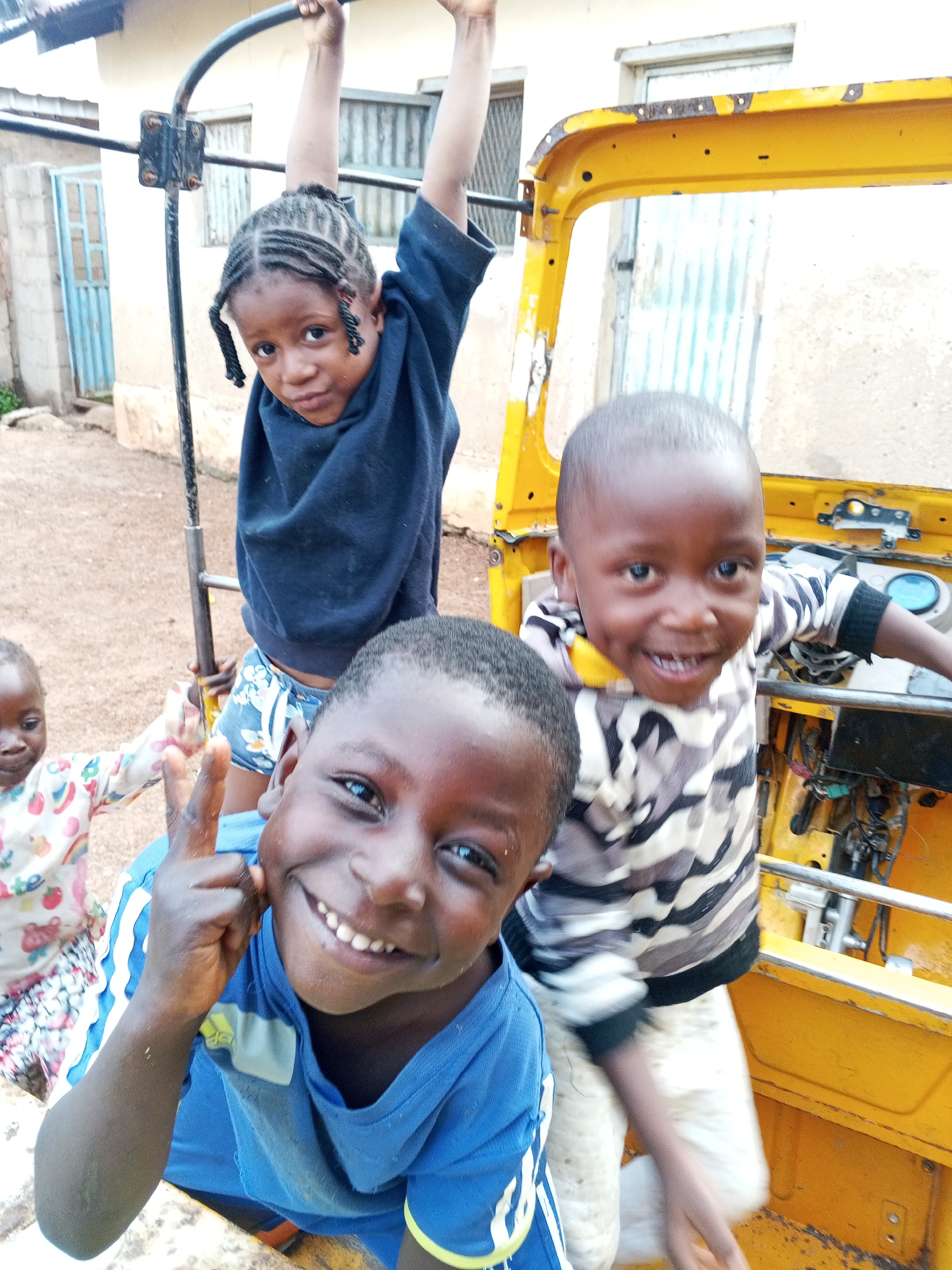 Four children are grouped around a yellow vehicle, possibly a dismantled auto rickshaw. One child is hanging from a metal bar behind the vehicle. Another child is giving a peace sign in the foreground, while another stands inside the vehicle. The fourth child is partially visible on the left side. The setting appears to be an outdoor residential area, with a building visible in the background.