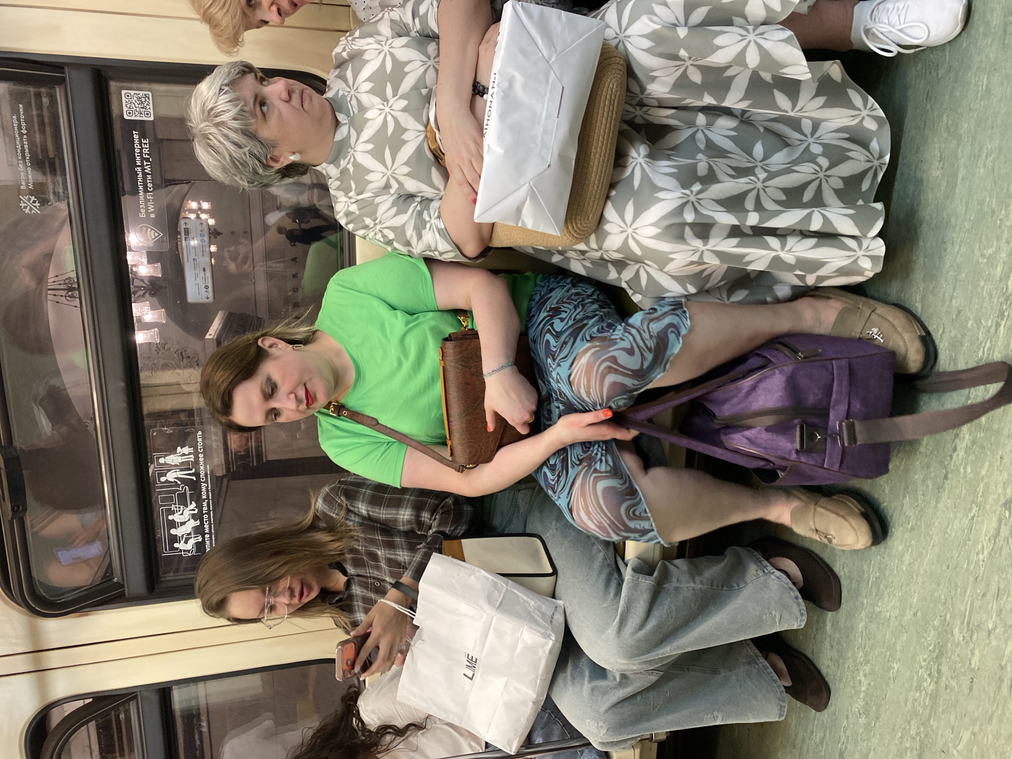 The image captures an interior scene within a Moscow Metro train in Russia. Three seated individuals are visible. From top to bottom:
1. A woman with short gray hair wears a gray and white floral dress and white sneakers, holding a white paper bag and a straw handbag.
2. A woman in a bright green t-shirt and a blue patterned skirt is sitting beside her, holding a brown handbag and a purple bag.
3. A woman with long brown hair wearing glasses and a plaid shirt is looking at her cell phone, with a white paper bag.
The metro window has signage. "Упите место тем, кому сложнее стоять" translates to "Give up your seat to those who have difficulty standing" and shows graphics depicting seated/standing people. "Вагон без кондиционера. Можно открывать форточки" translates to "Car without air conditioning. You can open the windows". The metro car also has "Безлимитный интернет в Wi-Fi сети МT\_FREE" which translates to "Unlimited internet in the MT\_FREE Wi-Fi network".