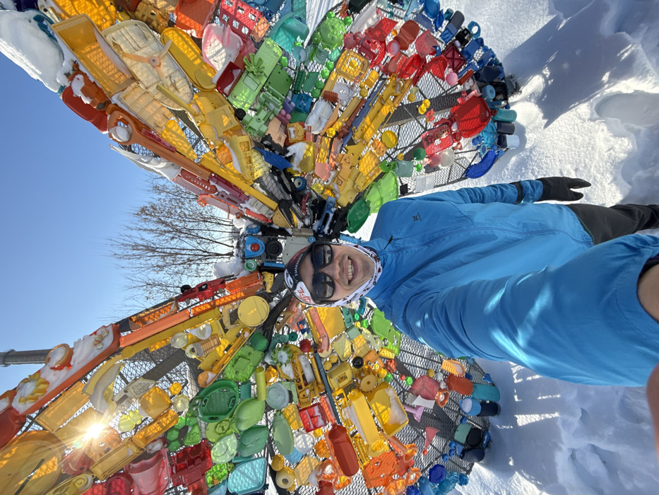 An adult male is captured in a selfie in front of a large, multicolored butterfly sculpture in Gorkinsko-Ometyevsky Forest Park, Kazan, Russia. The subject, positioned centrally, wears a bright blue zippered jacket, dark pants, black gloves, dark sunglasses, a white and red patterned neck gaiter, and a white beanie with "LIFE" text. He holds the camera, likely a smartphone, extended forward with his right hand and displays a smile. The butterfly sculpture, approximately life-size for an abstract design, is constructed from a dark metal grid frame adorned with numerous translucent and opaque plastic objects of various colors, including bottle caps, bottles, and containers, forming a mosaic across its wings. Patches of snow are visible on the upper surfaces of the sculpture. The ground is covered in deep white snow, exhibiting discernible footprints. The sky is clear and blue, with the sun's glare prominent in the upper left, casting defined shadows on the snow. Bare deciduous trees and a slender, distant tower are visible in the background, indicating a clear winter day.
