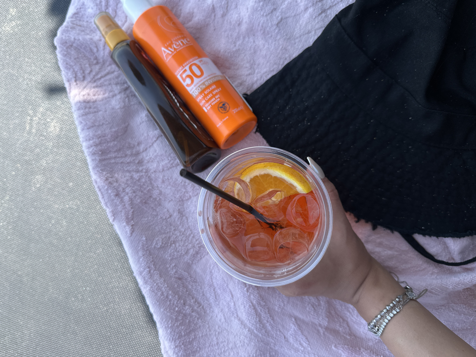 A hand wearing a silver bracelet holds a clear plastic cup filled with an orange-colored beverage with ice and an orange slice. The cup is on top of a pink towel. To the right of the cup are two bottles: a dark brown bottle and an orange bottle of Avène SPF 50 sunscreen. The pink towel is on top of a grey, textured surface, possibly a lounge chair. A piece of black fabric is visible at the bottom right of the image. The scene is set outdoors, likely at Ofrynio Beach, Greece, suggesting a relaxation or vacation setting.