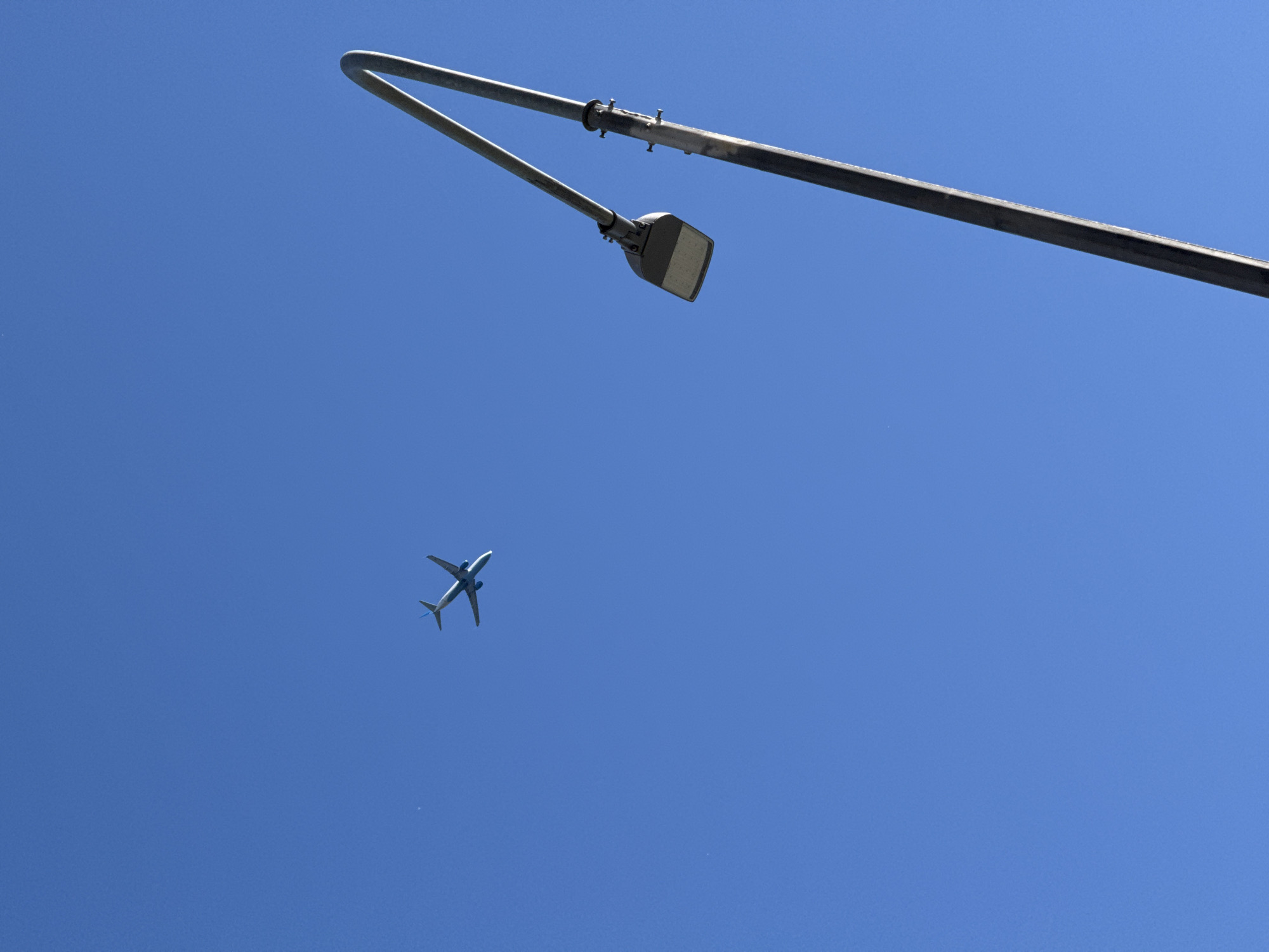 The media file features a clear, bright blue sky serving as the backdrop. Dominating the upper right portion of the frame is a street lamp post, made of metal, with its curved arm extending to the left and holding a modern, rectangular light fixture. Below and to the left of the lamp, a small, white airplane is visible, its wings extended and pointing upwards, indicating it is in flight. The airplane appears to be a commercial jet. The overall scene suggests daytime, with good visibility due to the clear sky. There are no visible people or text in the image. The location context of Saint Petersburg, Russia, suggests this could be an urban or suburban setting, with the street lamp being a common feature of such environments. The bright sky and clear air suggest fair weather.