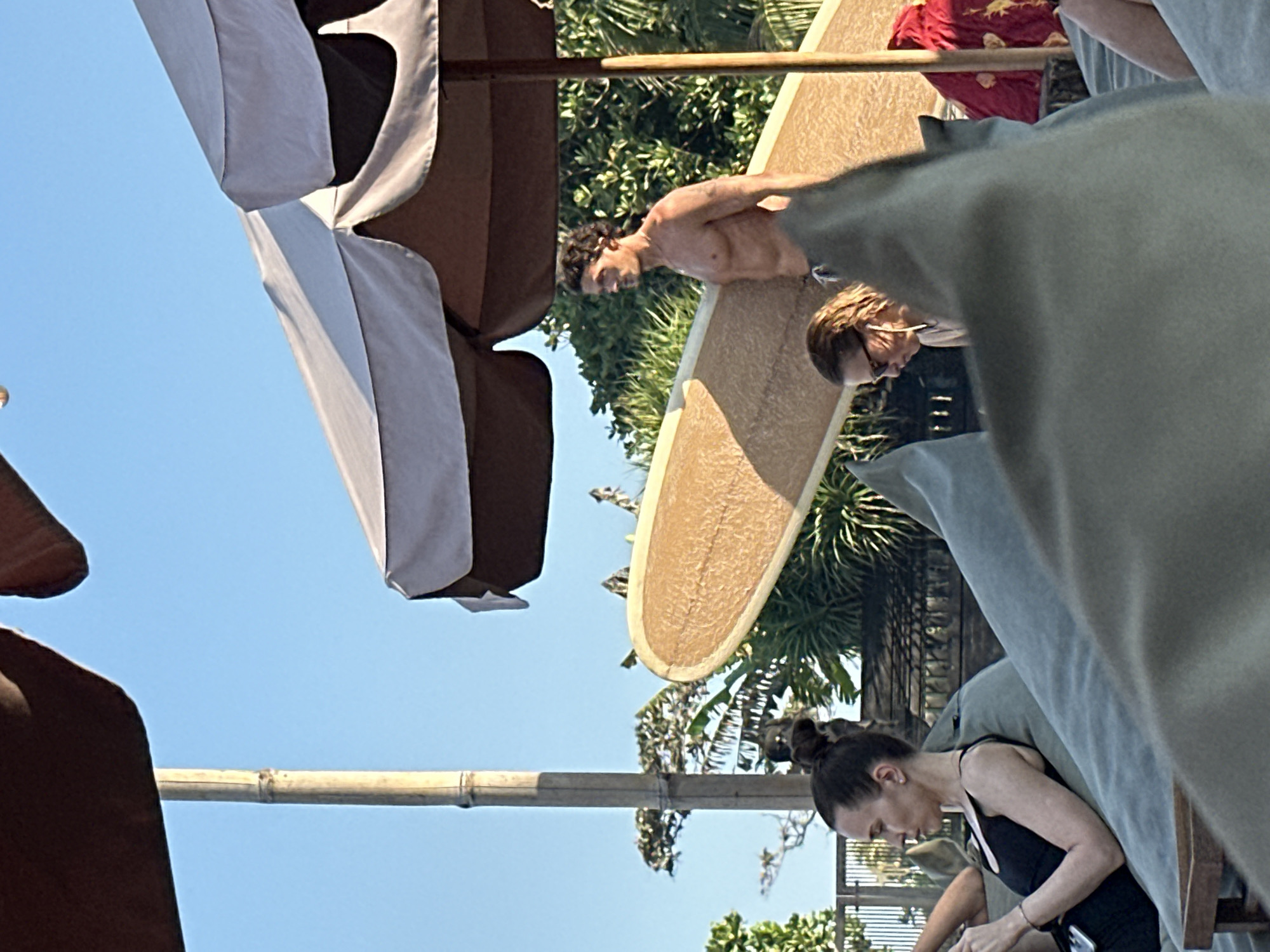 In North Kuta, Indonesia, a man with dark curly hair and a muscular physique, shirtless and wearing dark swim trunks, stands holding a light brown surfboard. He is partially obscured by a large, light-colored umbrella. In the foreground, a woman with her hair in a bun, wearing a black tank top, is seated on a large gray cushion, looking at her phone. To her right, another person wearing glasses, with light brown hair, is also seated on a cushion. The background features lush green foliage, a bamboo pole, and a fence. The scene appears to be outdoors, possibly at a beachside resort or cafe, under a clear blue sky.
