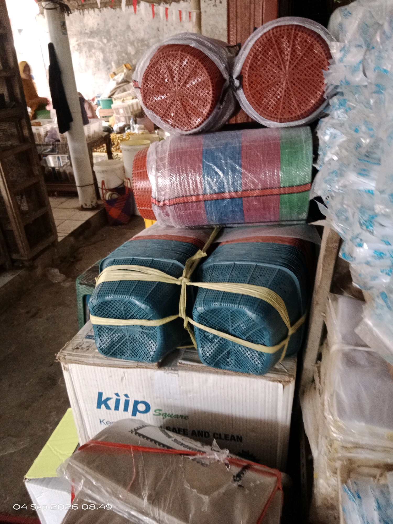The image shows a market stall in Kalipurwo, Indonesia, filled with various household items. In the foreground, there are stacks of blue plastic baskets tied with yellow rope, placed on top of a cardboard box. The box has blue lettering that reads "kiip Square" and further text including "SAFE AND CLEAN". Above the baskets, there are two red circular plastic items wrapped in plastic, possibly strainers or colanders, stacked on top of a roll of striped matting. To the right, a stack of clear plastic bags filled with white items is visible. In the background, there are glimpses of other market stalls with goods, a person wearing a yellow headscarf, and a white pole. The overall environment appears to be an indoor market with a rough concrete floor and a partially visible corrugated ceiling with red and white decorative flags. The lighting suggests it is daytime.