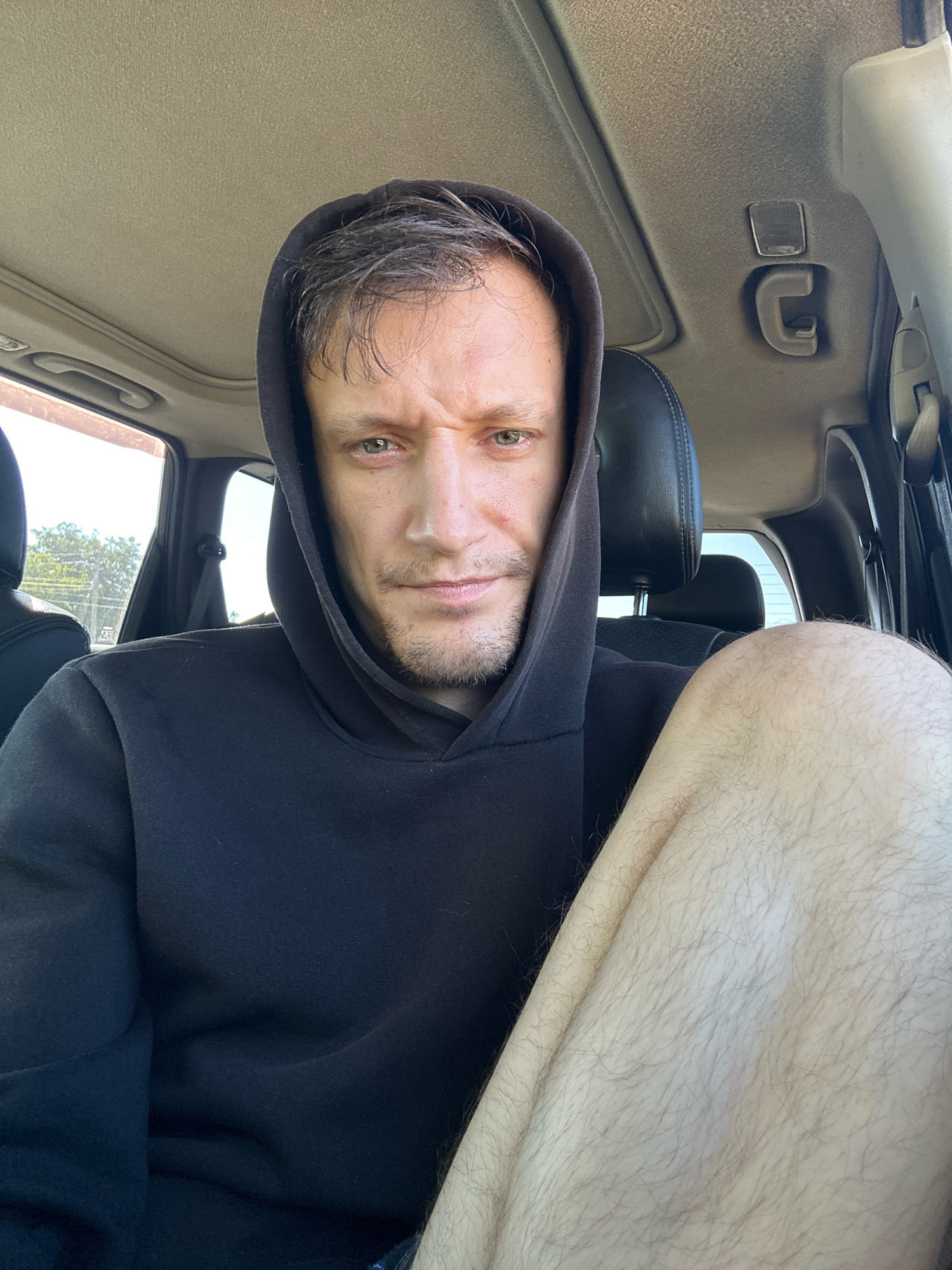 A man wearing a black hoodie sits in the driver's seat of a car, looking directly at the camera with a furrowed brow. His right leg, clad in denim shorts and covered in light brown hair, is prominently displayed in the foreground. The car's interior is visible, including the sunroof, headliner, and part of the passenger seat. The background suggests an outdoor environment, possibly a street or parking lot, with trees and a fence visible through the car window. The overall impression is that of a casual, informal portrait taken from inside a vehicle. The location of Makiv, Ukraine is not discernible from the visual information provided in the image.