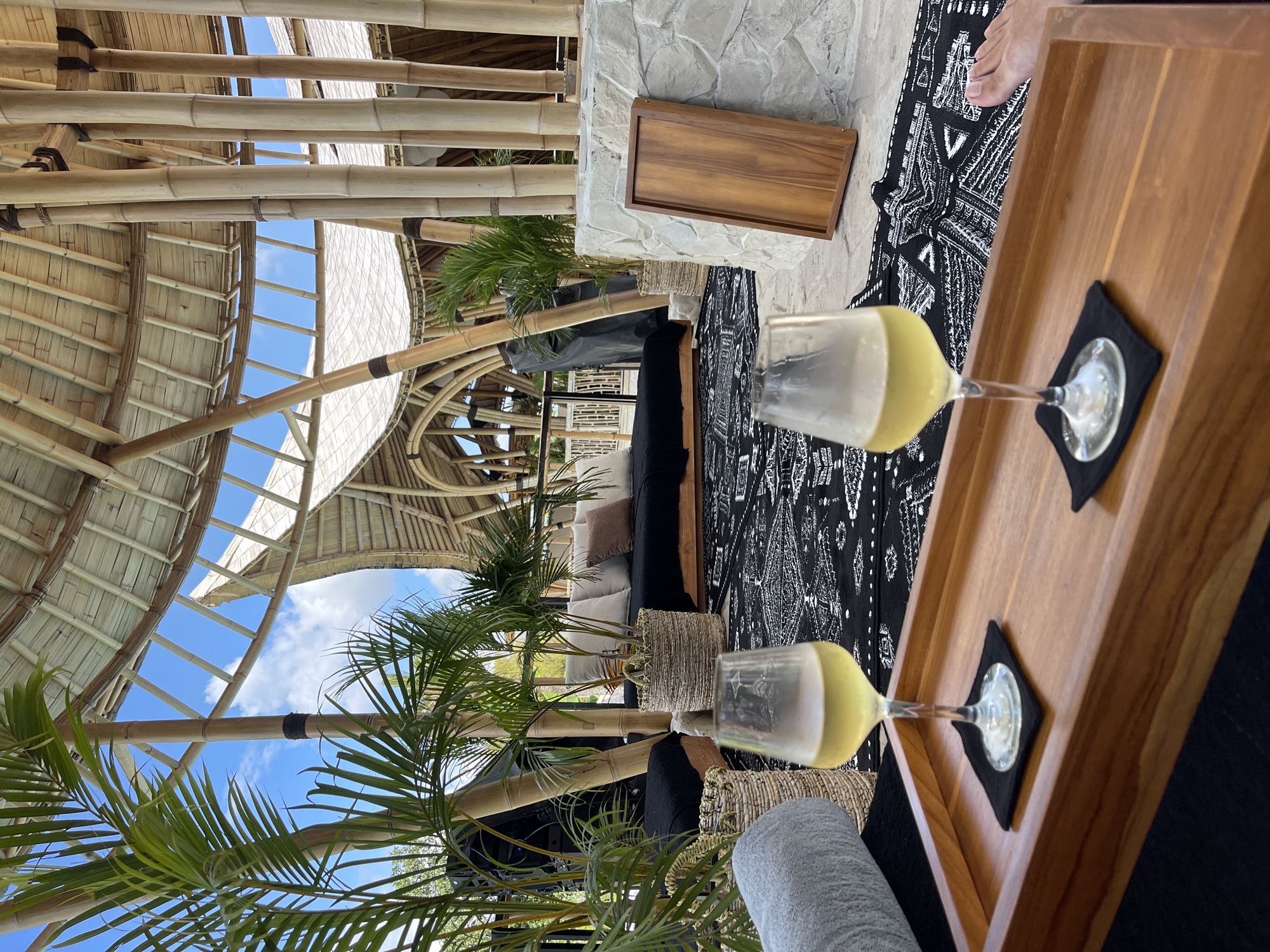 A high-angle shot captures two glasses of white wine resting on a wooden tray placed on a black and white patterned rug. The glasses are in the foreground, with the tray extending into the middle ground. Behind the tray, a black couch with white and brown pillows is visible, accompanied by potted plants and woven baskets. The scene is set within an open-air structure with a bamboo ceiling and columns, suggesting an outdoor dining or lounging area. The environment appears to be a resort or a high-end establishment, possibly in Ubud, Indonesia, given the architectural style and tropical vegetation. The sky is visible through gaps in the bamboo ceiling, indicating a clear day. A portion of a person's foot is visible on the right side of the image.