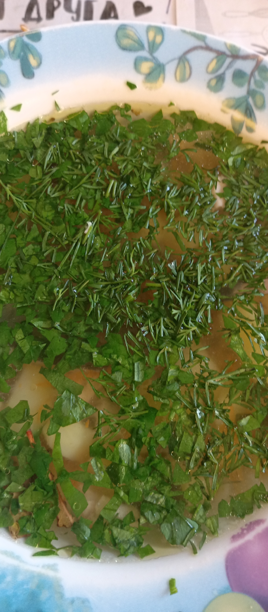 A close-up shot presents a bowl of soup, generously garnished with freshly chopped herbs, predominantly dill and parsley. The soup itself appears to be a clear broth, possibly chicken or vegetable-based, with visible chunks of potato and other ingredients submerged within. The bowl is decorated with a blue and white floral pattern around the rim. Part of a word in a different language, possibly "ДРУГА," and a heart symbol are visible above the bowl. The scene suggests a home setting. No city or location is known.