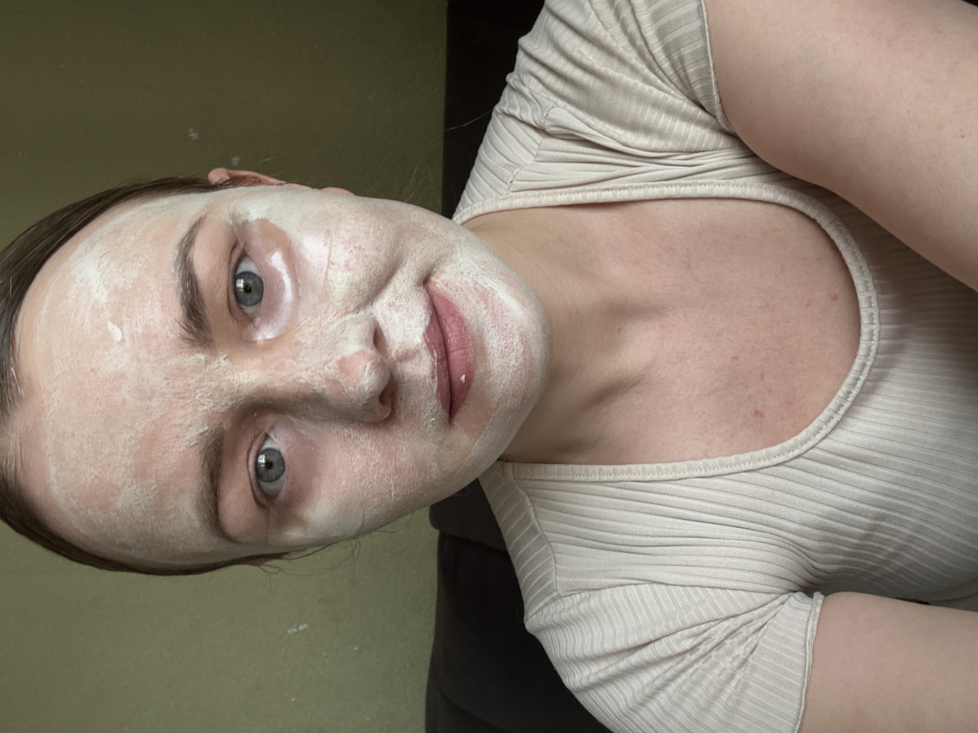 A woman with fair skin and light blue eyes is shown close-up, facing the camera at an angle. She has a white face mask applied to her face, which covers her forehead, cheeks, nose, and chin but stops around her lips. She is wearing a beige ribbed shirt with a round neckline. The background is a solid pale green color in the top left, while the rest of the background is dark and out of focus. It is impossible to tell from the information provided in the image alone if this image was taken in Chaikovsky, Russia.