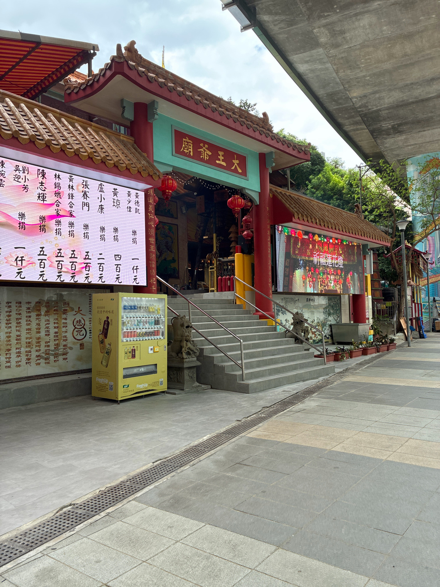 This image captures an outdoor scene in front of a traditional East Asian temple, likely in Hong Kong, China, given the architectural style and visible text. The temple features a red and green facade with intricate tiled roofs, characteristic of Chinese temple architecture. In the foreground, a bright yellow vending machine stands on a paved walkway. A set of concrete stairs leads up to the temple entrance, flanked by metal railings. On the left side of the temple, a large digital billboard displays colorful text and images. Another digital screen is mounted above the temple entrance, showing a vibrant image of lanterns. The environment appears to be urban, with a concrete overpass visible in the upper right portion of the image, suggesting the temple is located near a modern infrastructure. The sky is partly cloudy. There are no people visible in the immediate vicinity of the temple or the vending machine, giving the scene a quiet atmosphere.