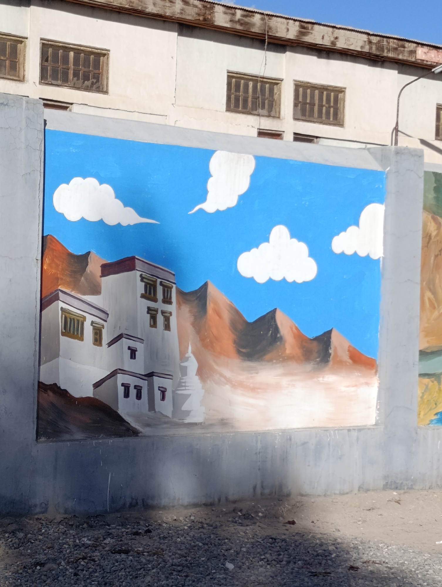 The image shows a mural painted on a grey concrete wall in Leh, India. The mural depicts a landscape with a bright blue sky filled with white, puffy clouds. In the foreground of the mural, there are white buildings with dark windows, resembling traditional Ladakhi architecture. Behind the buildings, stylized mountains with reddish-brown and grey hues rise up. The mural appears to be on an exterior wall, possibly part of a building or a boundary wall, with gravel on the ground in front of it. Above the mural, the upper part of a cream-colored building with several barred windows can be seen, and a rusted metal structure is visible at the very top. A curved metal pole, possibly for a street light, extends upwards on the right side of the image. The overall scene suggests an outdoor urban or semi-urban environment.