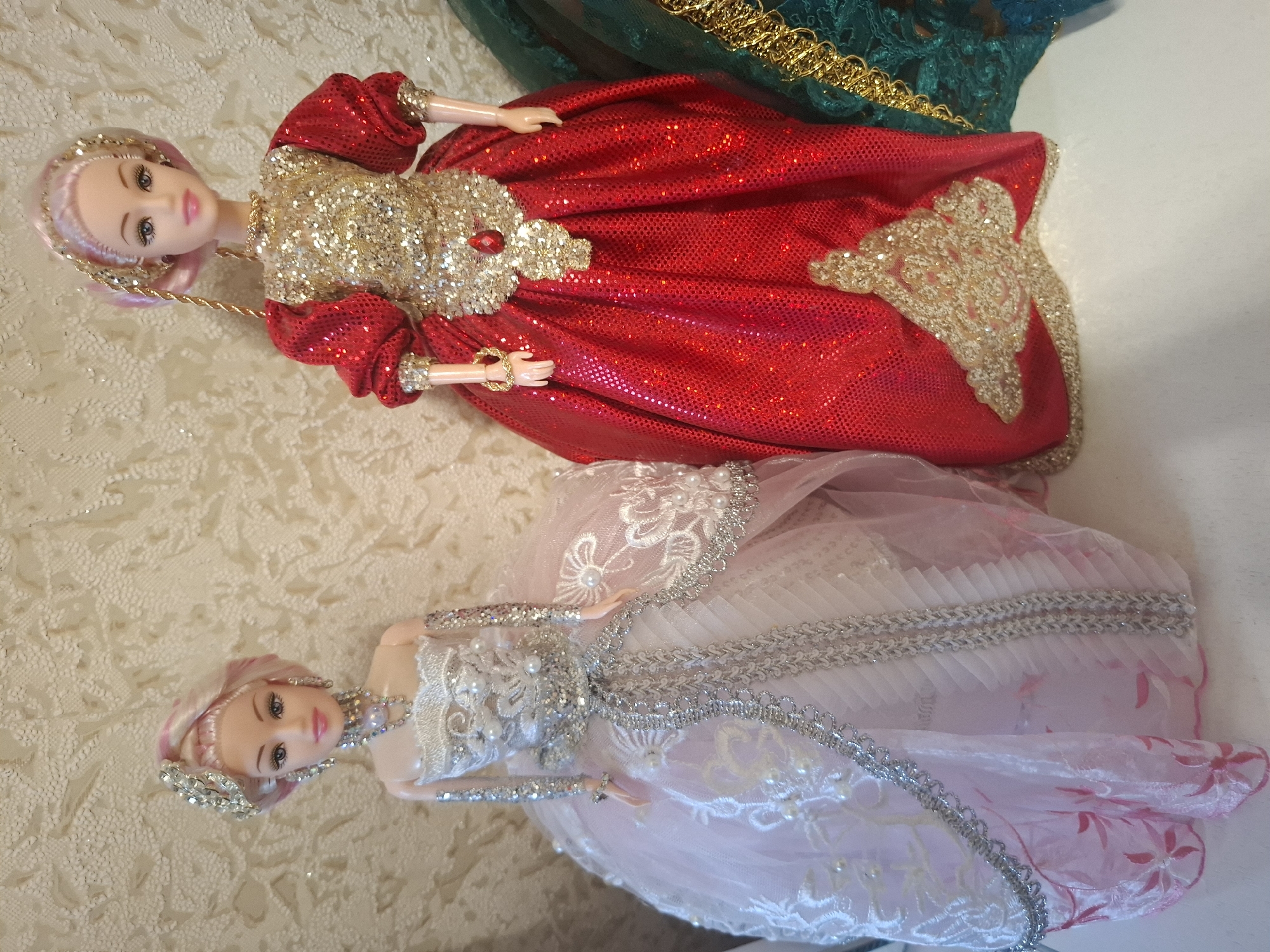 Two dolls are shown lying on a textured, cream-colored surface. The doll in the foreground is wearing a pink dress with white lace and silver trim. She has blonde hair with pink streaks and is wearing a silver headband and necklace. The second doll, behind the first, is wearing a sparkly red dress with gold embellishments and a red jewel on the chest. She has blonde hair with pink streaks and is wearing a gold headband. The dolls are posed as if standing, with their arms slightly bent. The background is a busy, textured fabric that appears to be lace or a similar material. The lighting is bright, suggesting an indoor setting with artificial light. There is no visible text in the image. The dolls appear to be decorative, possibly part of a collection or display.