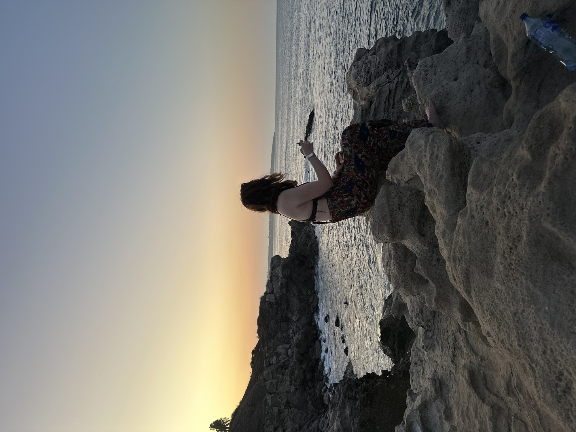 A woman sits on a rocky outcrop overlooking the ocean at sunset. She wears a dark bikini top and a patterned skirt. She holds a dark object in her hand. The water is visible in the background, with a bottle of water sitting on the rocks next to her. The location is likely near Puerto Escondido, Mexico, based on the coastal rock formations and sunset hues.