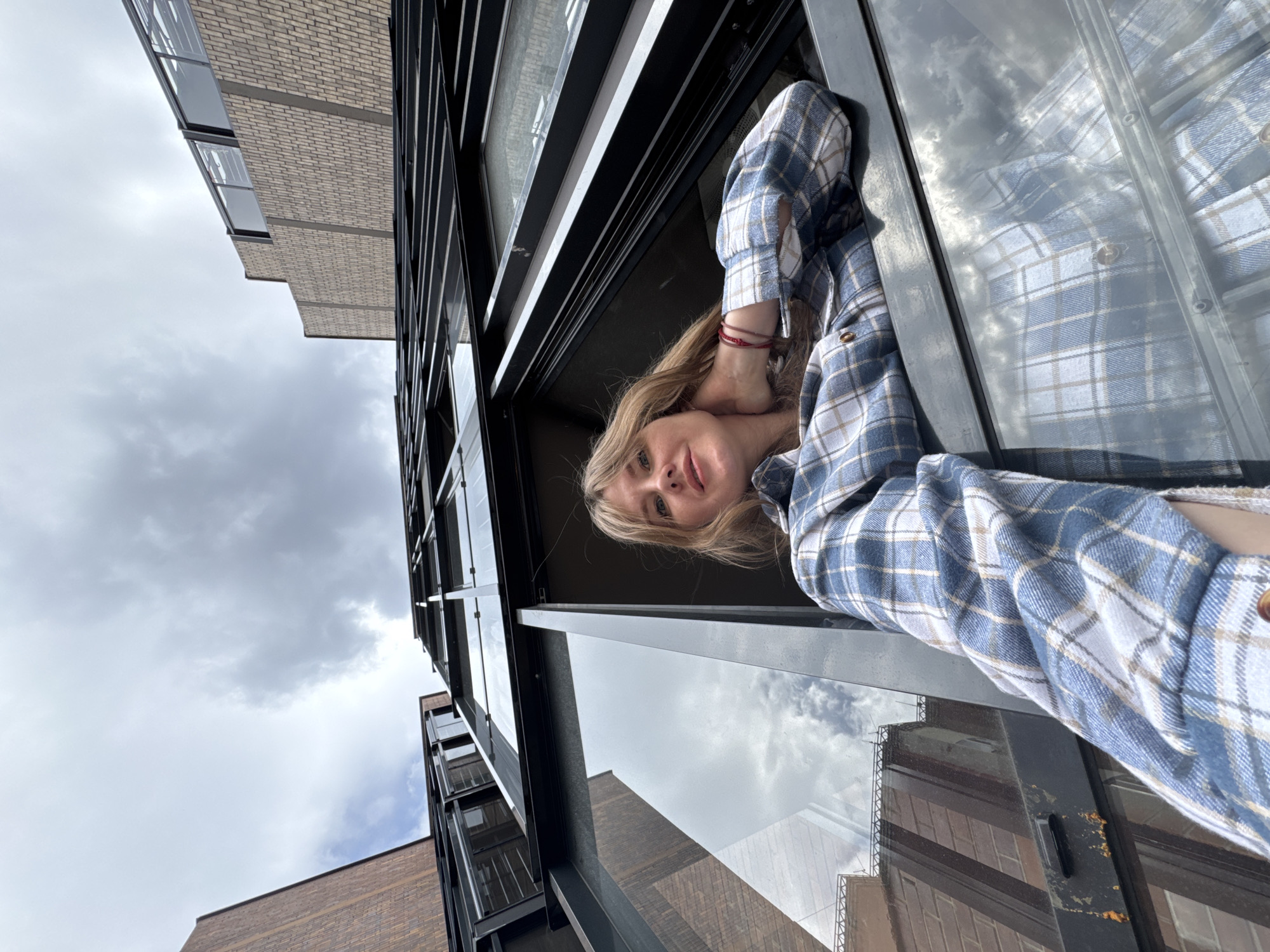 This low-angle, horizontally oriented image captures a young woman leaning out from a window or balcony of a modern building in Perm, Russia, set against a cloudy daytime sky. The woman, with blonde hair, wears a blue, white, and tan plaid flannel shirt. Her left arm is bent with her hand near her neck, and a red bracelet is visible on her wrist. She gazes directly at the viewer with a subtle smile.
The surrounding architecture is characterized by dark metal window frames and light-colored brick walls, some with a prominent horizontal pattern. The expansive cloudy sky, indicative of an overcast day, fills a significant portion of the background. Reflections of the sky and other nearby structures are evident on the building's glass surfaces, adding depth to the scene. The unconventional camera angle places the woman horizontally across the frame, creating a dynamic and somewhat disorienting perspective. No visible text is present in the image.