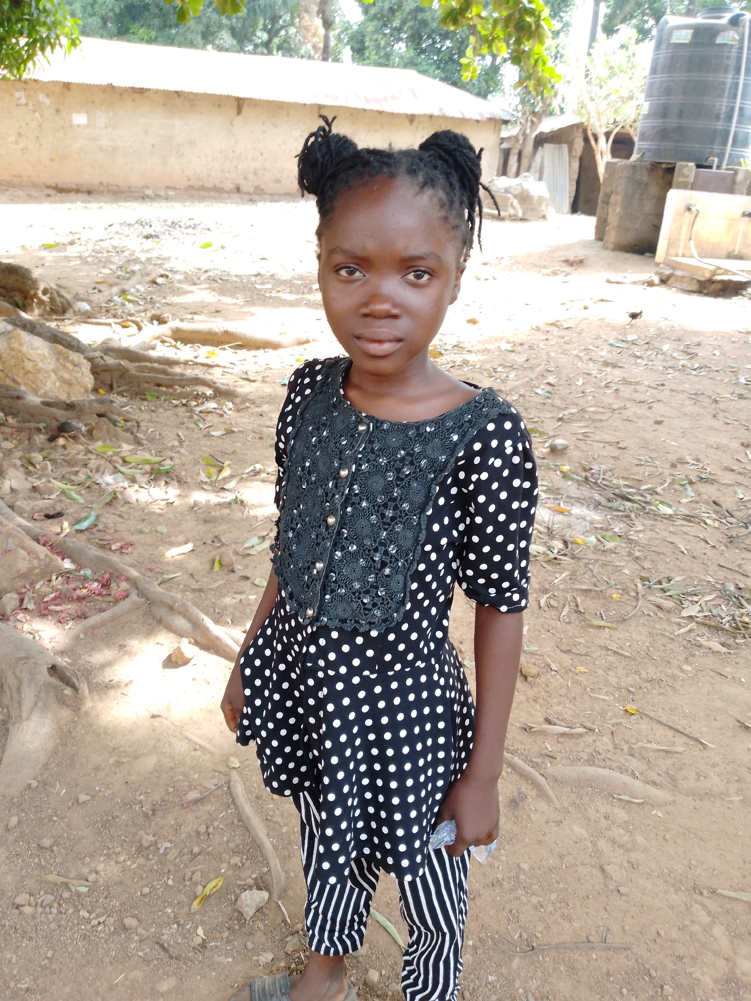 A young girl with braided hair stands outdoors on dirt ground in Akwanga, Nigeria. She wears a black and white polka dot top with lace detailing and striped pants. Behind her is a building and various outdoor objects, including a water tank.
