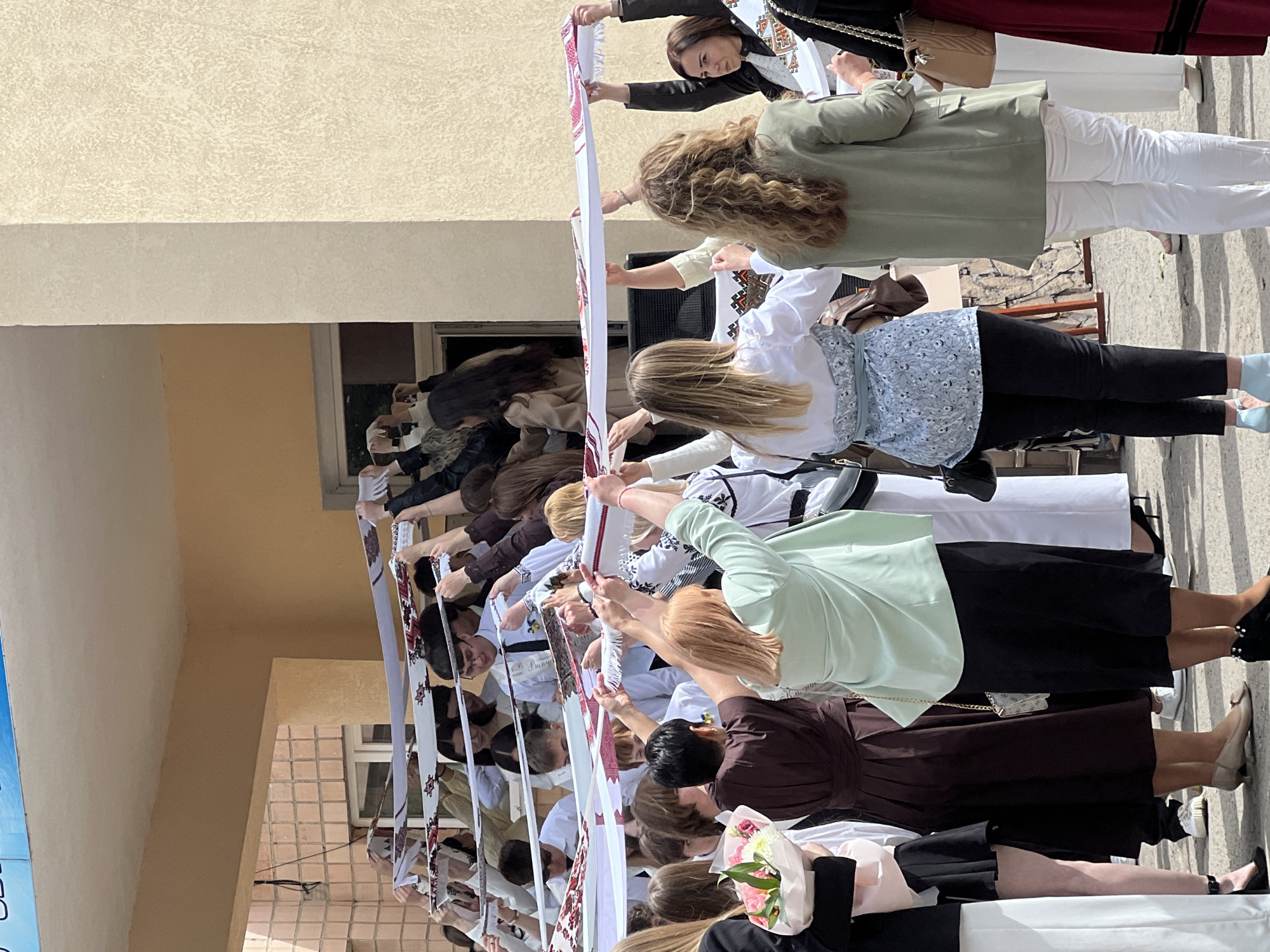 A crowd of people are holding up a long, white cloth strip with red embroidered designs on it. The photo appears to have been taken in Ternopil, Ukraine, based on the architecture and possibly the embroidered designs on the cloth. Most of the people in the crowd are young adults, possibly students, dressed in a variety of casual attire. The setting appears to be an outdoor area next to a building, as evidenced by the wall and windows visible in the background.