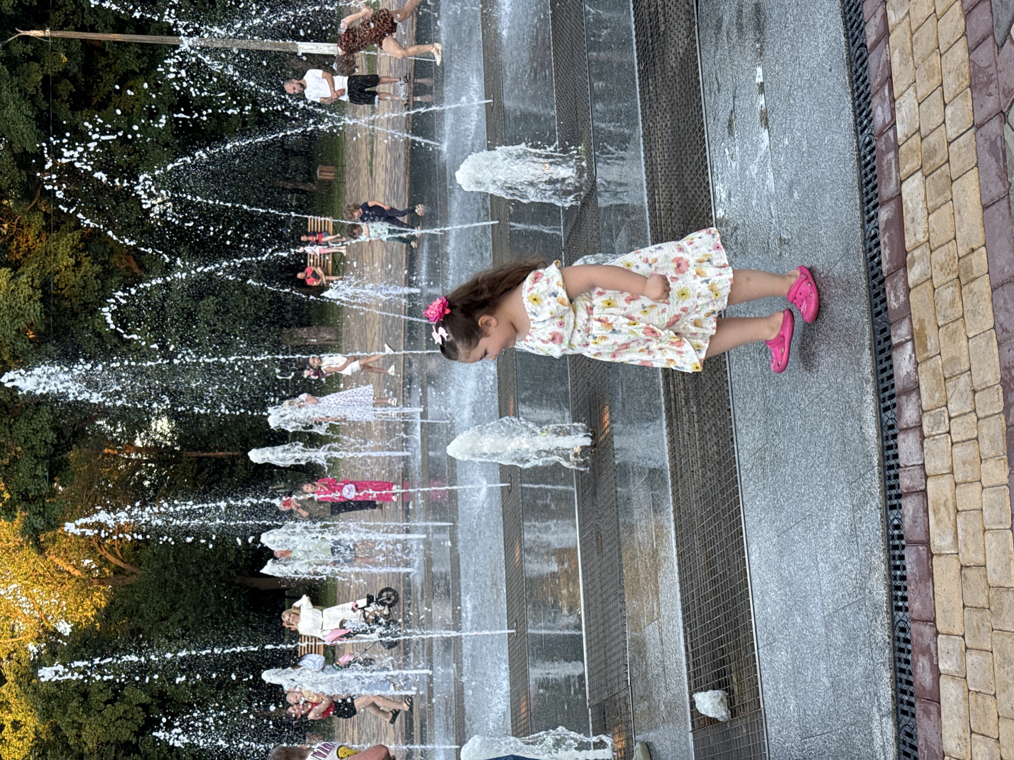 A girl in a floral dress and pink shoes stands by a fountain. She is looking to her left. In the background, other people, including children and adults, are visible interacting with the fountain. The fountain has multiple streams of water shooting upwards. The area around the fountain is paved with grey stone and metal grates. The background consists of trees and a lamppost.