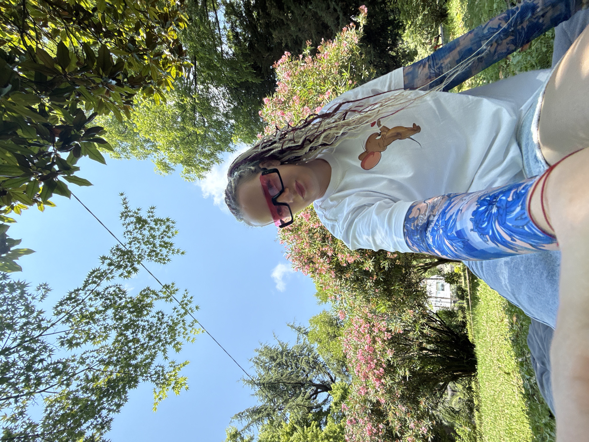 The image shows a young woman with blonde and dark braided hair, wearing large pink-tinted sunglasses and a white t-shirt with a cartoon mouse graphic. She is outside, in what appears to be a park or garden setting. Lush green trees and bushes with pink flowers are visible in the background, along with a bright blue sky. The woman's arms are adorned with blue and white patterned sleeves. The photo seems to be a selfie, taken from a low angle looking up towards the sky and the woman. The sunlight suggests it is daytime. The presence of green foliage and blooming flowers indicates a warm season. The overall impression is one of a relaxed, casual outdoor moment.