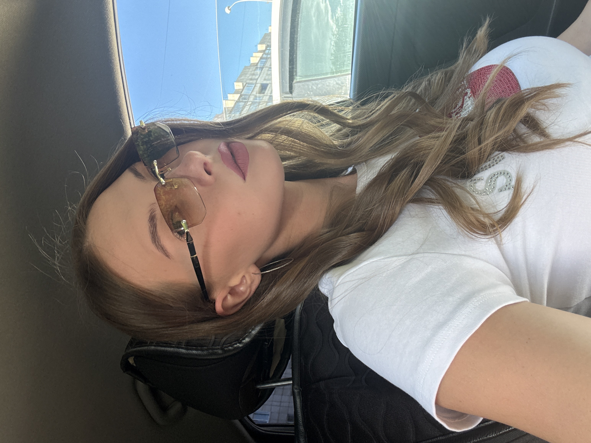 The image shows a young woman sitting in the driver's seat of a car, with her head tilted to the side, looking away from the camera. She is wearing sunglasses, a white t-shirt with a design on the chest, and a thin hoop earring in her left ear. The background shows buildings and a clear blue sky, suggesting an outdoor urban environment. There is no discernible indication of the city of Perm, Russia, or any other specific location. The car interior is visible, including the headrest of the passenger seat. The lighting suggests it is daytime.