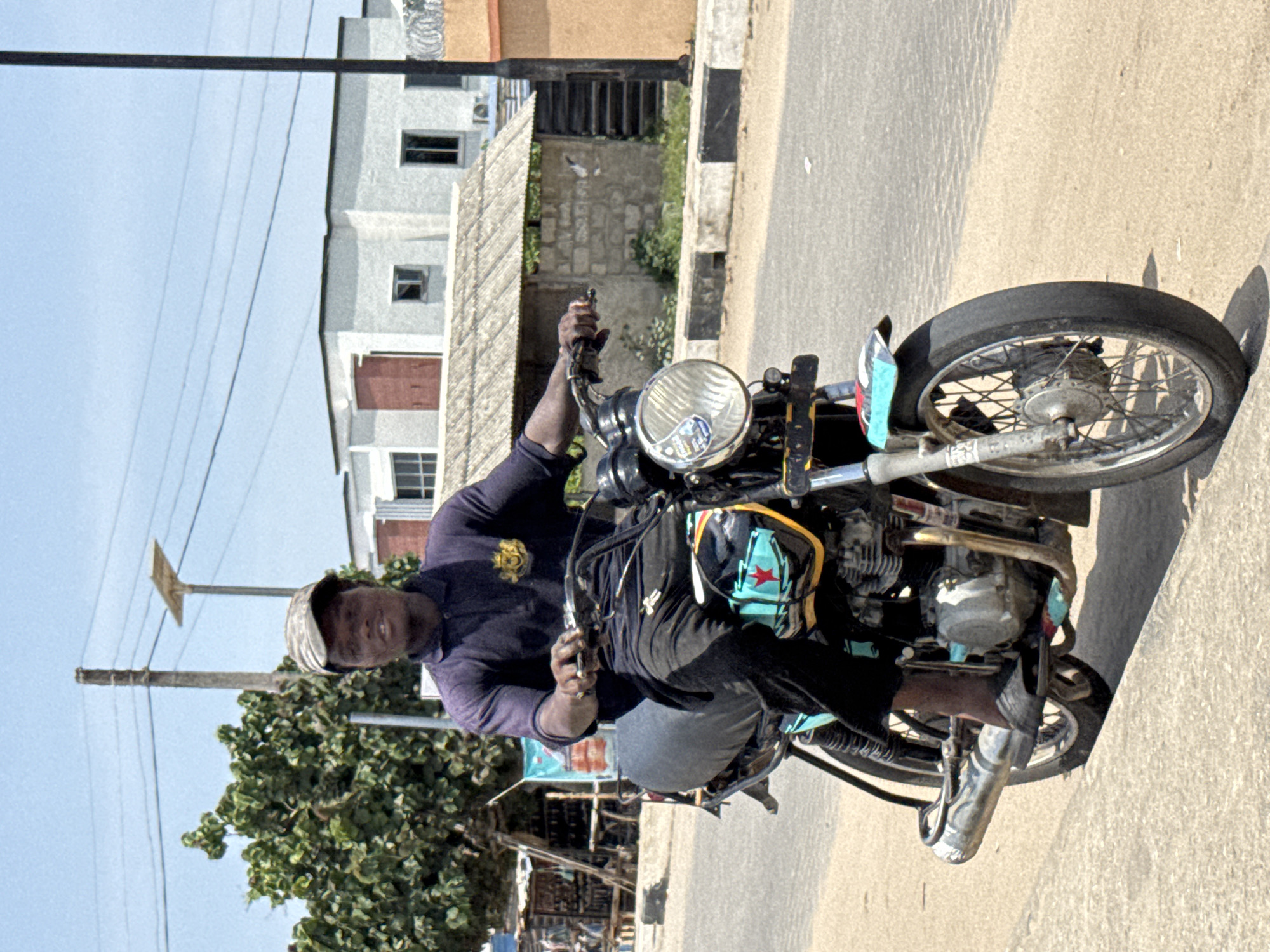 A man is riding a motorcycle on a paved road in Badagry, Nigeria. He is wearing a dark-colored long-sleeved shirt and a hat. The motorcycle is an older model, and a bag with a red star is strapped to the front. Buildings and trees are visible in the background, along with utility poles and wires. The sky is clear and blue.