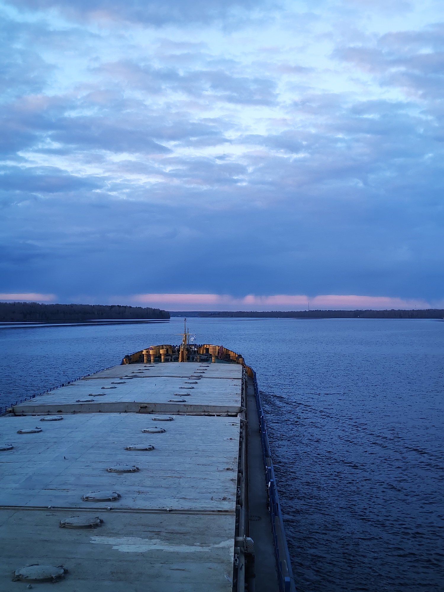 The image depicts a barge traveling on a river, likely the Volga River near Turtsevo, Russia. The barge deck is visible in the foreground, with numerous hatches. The water surface is slightly rippled. In the distance, the riverbanks are lined with trees and shrubbery. The sky is overcast with a mixture of blue and gray clouds, suggesting either sunrise or sunset. A faint orange hue is visible near the horizon line. A tall, thin structure (possibly a radio tower) is visible on the distant right riverbank. No people are visible.