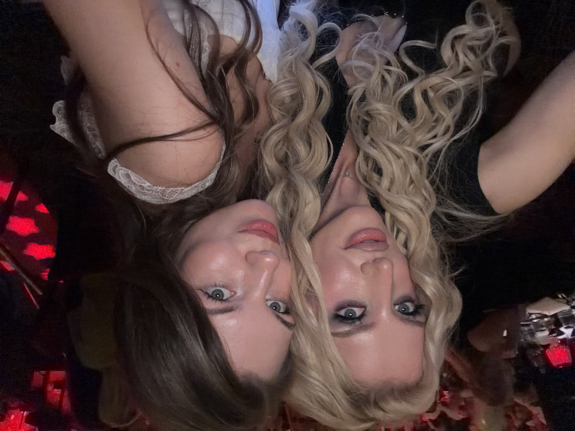 Two Caucasian women are close together, posing in a selfie. The woman on the left has long brown hair and green eyes, and is wearing a white top with lace trim. The woman on the right has blonde, curly hair and blue eyes, and is wearing a black top. The image appears to have been taken in a dimly lit location, possibly a nightclub or bar, given the red lighting and the presence of glasses in the background. The location of the city of Belgrade, Serbia is not apparent from the provided context.