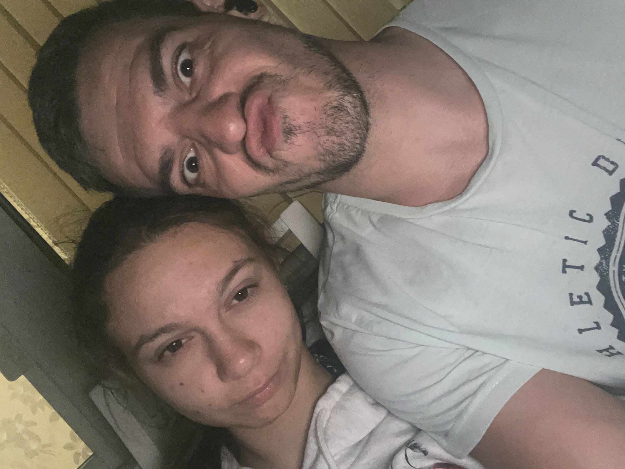 A close-up selfie shows a couple, a woman and a man, lying down. The woman is on the left, her head tilted towards the man, with her eyes open and a slight smile. The man is on the right, making a funny face with his lips puckered and eyes wide open. Both are wearing casual t-shirts. The background appears to be indoors, possibly a bedroom, with wooden paneling and a light-colored wall with a floral pattern. The lighting suggests it is nighttime or a dimly lit indoor space. The image does not contain any visual information that could place it in Sofia, Bulgaria.