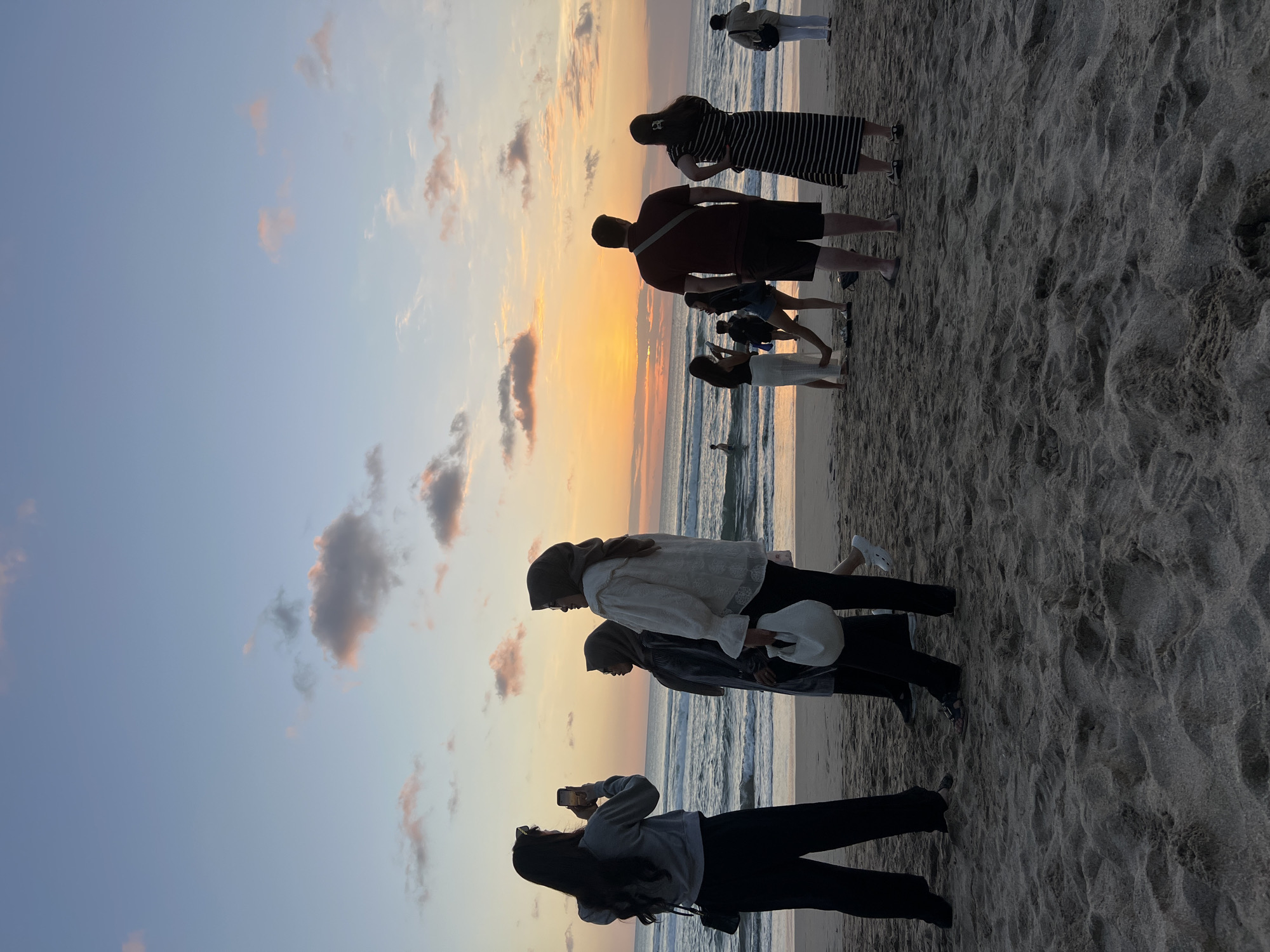 A group of people are standing on a sandy beach in Kuta, Indonesia, during sunset. The sky is a gradient of soft blues and oranges, with scattered clouds. The ocean waves are gently rolling onto the shore. In the foreground, a woman on the left is taking a picture with her phone. To her right, two women are standing, one in a white long-sleeved top and headscarf, and the other in a dark grey headscarf and dress. Further to the right, a man in a maroon t-shirt and shorts is facing away from the camera, and a woman in a black and white striped dress is beside him. Other individuals and groups are visible in the background, some wading in the water. The overall scene depicts a peaceful evening at the beach.