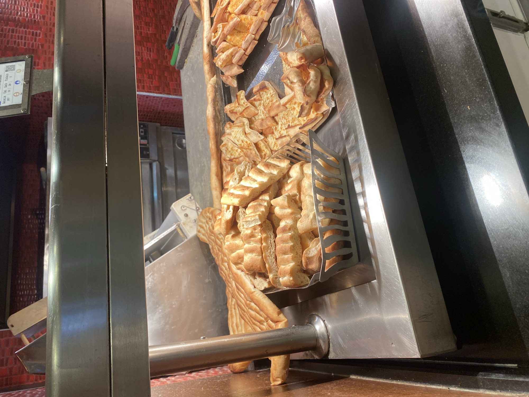 The image shows a buffet display of baked goods, likely bread and flatbreads, at a food establishment in Aksu, Turkey. The items are arranged on stainless steel surfaces under a warm light. In the foreground, a silver basket contains cut pieces of bread. To the right of the basket, a tray holds triangular slices of flatbread topped with melted cheese. Further back, larger, whole flatbreads with scored patterns are visible. The background includes a red tiled wall and a commercial oven. The overall scene suggests a self-service food area in a bakery or restaurant.