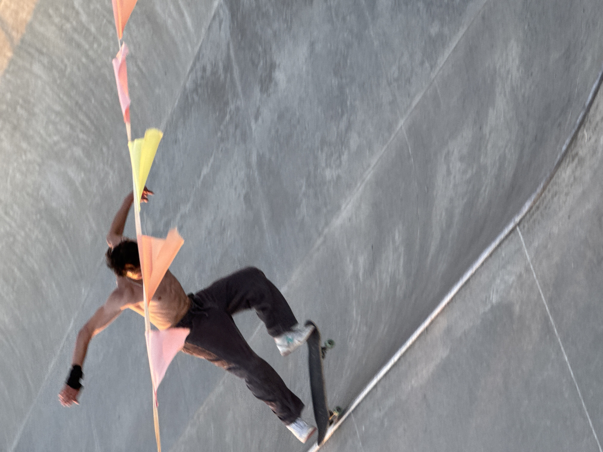 A shirtless male skateboarder in dark pants and white socks is captured in motion within a concrete skatepark. He is wearing a black wristband on his left arm and his body is angled, suggesting he is in the midst of a trick or maneuver. His right leg is bent, and his left leg is extended, positioned over a black skateboard. The skateboard has green wheels. A string of colorful flags, predominantly pink and yellow, is strung across the upper portion of the image, appearing to be decorative elements within the skatepark. The skatepark itself is constructed of grey concrete with visible lines and seams, indicating a contoured surface. The lighting suggests it is either sunrise or sunset, casting a warm glow on the scene. The location is identified as Sochi, Russia.