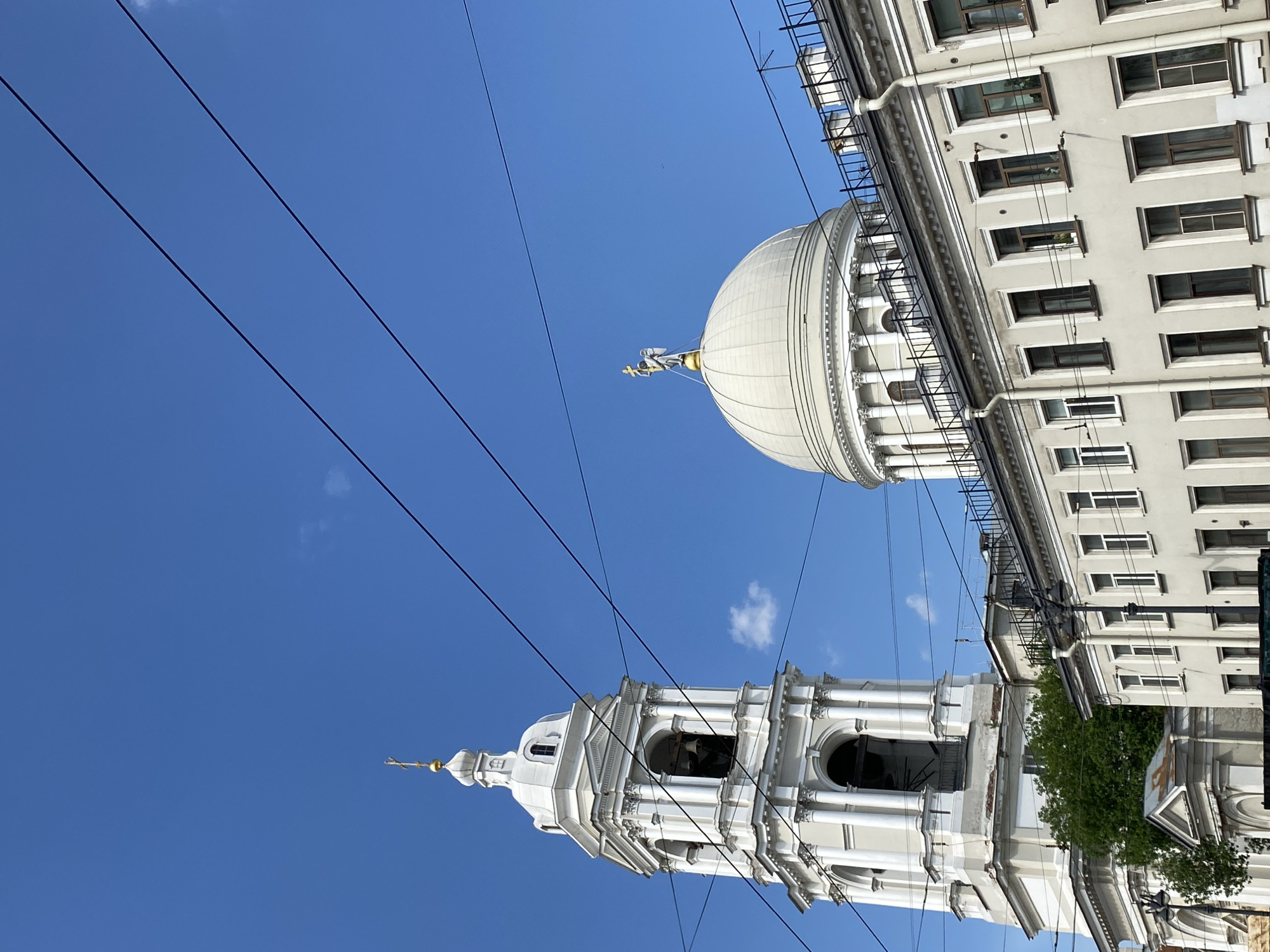 This vertical, eye-level shot captures architectural elements against a clear blue sky, likely in Saint Petersburg, Russia, based on the provided context. The image is oriented such that the sky occupies the upper and left portions, while buildings are on the right and lower parts.
On the left, a grand, white architectural structure rises, resembling a church bell tower or belfry. It features multiple tiers with ornate classical detailing, including columns, arched openings (likely for bells), and a domed top culminating in a golden finial. To its right, and slightly higher in the frame, is a large, pristine white dome, also part of a significant building. This dome is topped with a golden statue, possibly depicting an angel or a classical figure.
Further to the right, a multi-story, light-colored building with numerous rectangular windows is partially visible, suggesting a more residential or commercial urban structure. A few patches of green foliage from trees or bushes are visible near the base of these structures, indicating street level.
Several dark overhead wires, likely for public transport like trams or trolleybuses, crisscross prominently against the bright sky, adding a modern urban element to the historical architecture. The weather is sunny with a clear blue sky and only a few faint, wispy clouds visible. The bright lighting suggests it is daytime. No discernible text is visible on any of the structures.