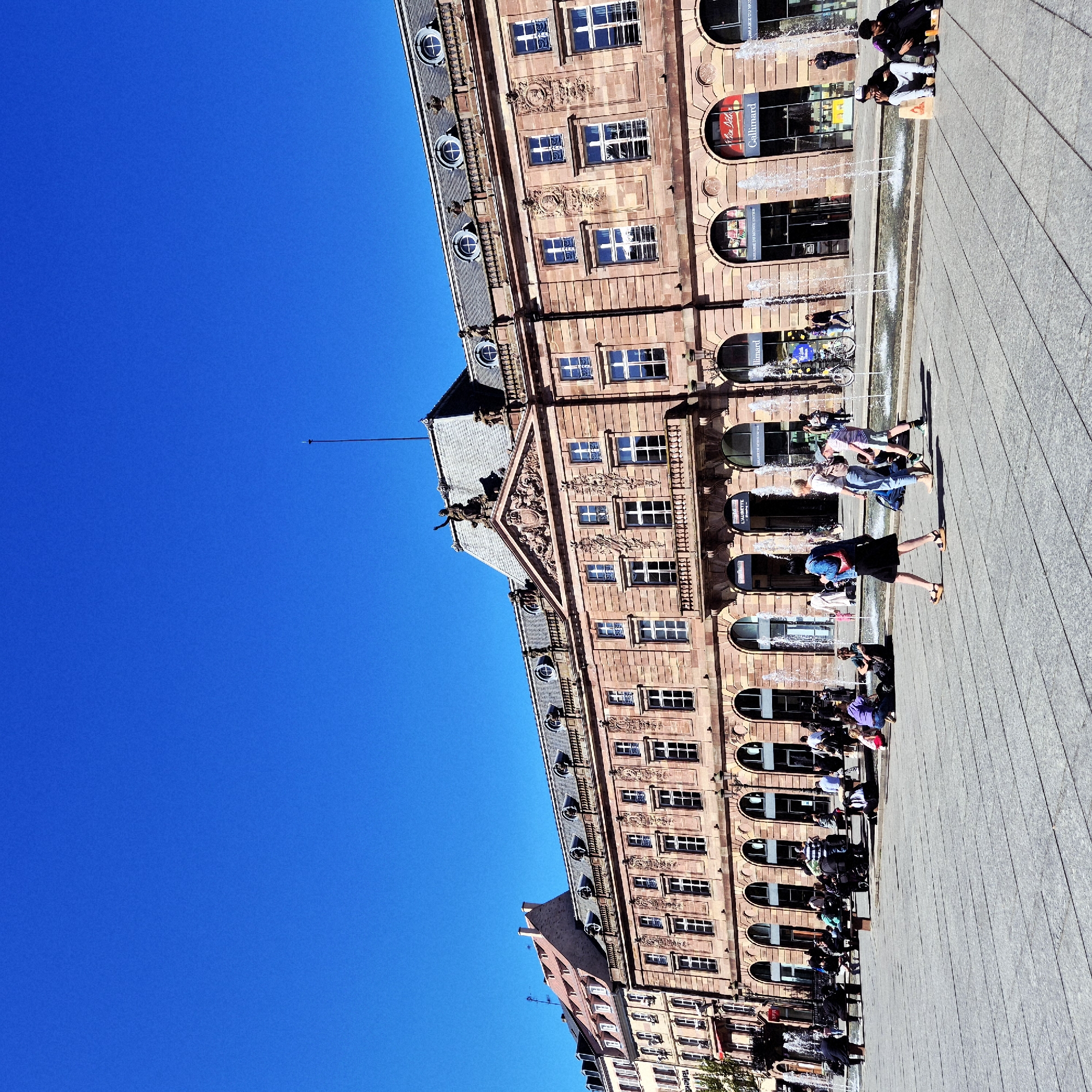 The image captures a view of Strasbourg, France. The iconic architecture is visible against a backdrop of a clear blue sky. The foreground shows a paved public space where people are engaged in various activities, including walking, sitting, and gathering near what appears to be a water feature or fountain. The building's architectural details, such as windows and decorative elements, are discernable. The people are dressed in a range of casual attire, indicating a relaxed atmosphere.
