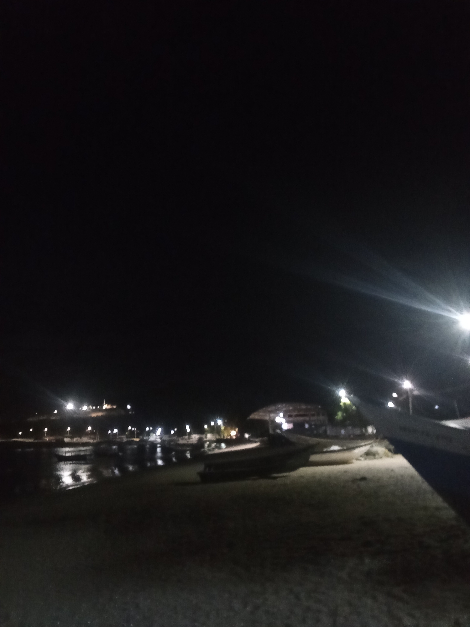 The image captures a night scene on the beach in Juan Griego, Venezuela. The scene is dominated by darkness, with only a few sources of light illuminating the landscape. In the distance, there's a building lit up, possibly a hotel or landmark, atop a hill. Closer to the viewer, there are boats on the shore, some near the water's edge. A streetlamp or other bright light source is visible on the right side of the image, causing glare. The ground is sandy, indicating the beach setting.