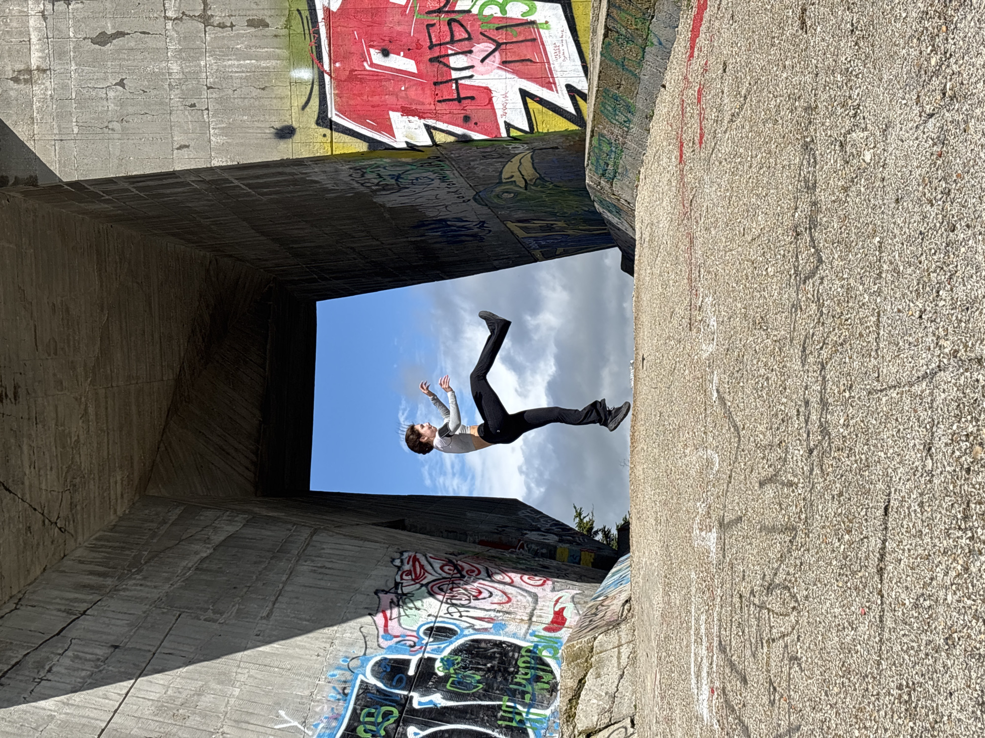 The photograph shows a person leaping into the air inside a concrete structure covered in graffiti. The person has dark hair and is wearing a light-colored long-sleeved shirt and dark pants, with sneakers. The person is in mid-air, with arms raised above the head and legs bent at the knees. The structure appears to be part of a concrete tunnel or bunker-like structure. The walls are covered with graffiti in various colors. Above the structure, a partly cloudy sky is visible. I am unable to determine whether the image was taken in Varna, Bulgaria based on the visual information provided.