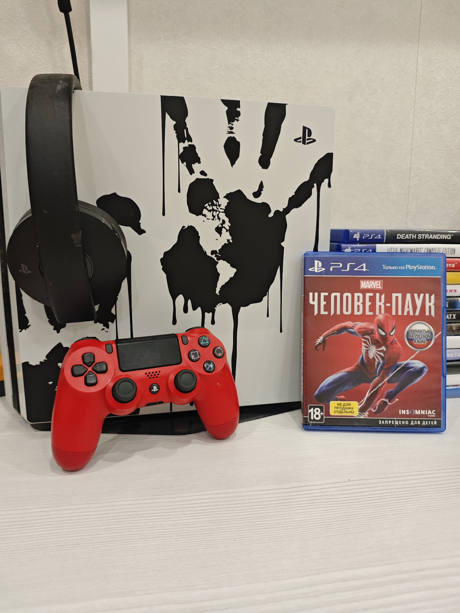 The image shows a PlayStation 5 console with a black and white design, a red PlayStation 4 controller, a black headset, and a stack of PlayStation 4 games, including a copy of "Marvel's Spider-Man" in Russian. The scene is set on a white, possibly wooden, surface.