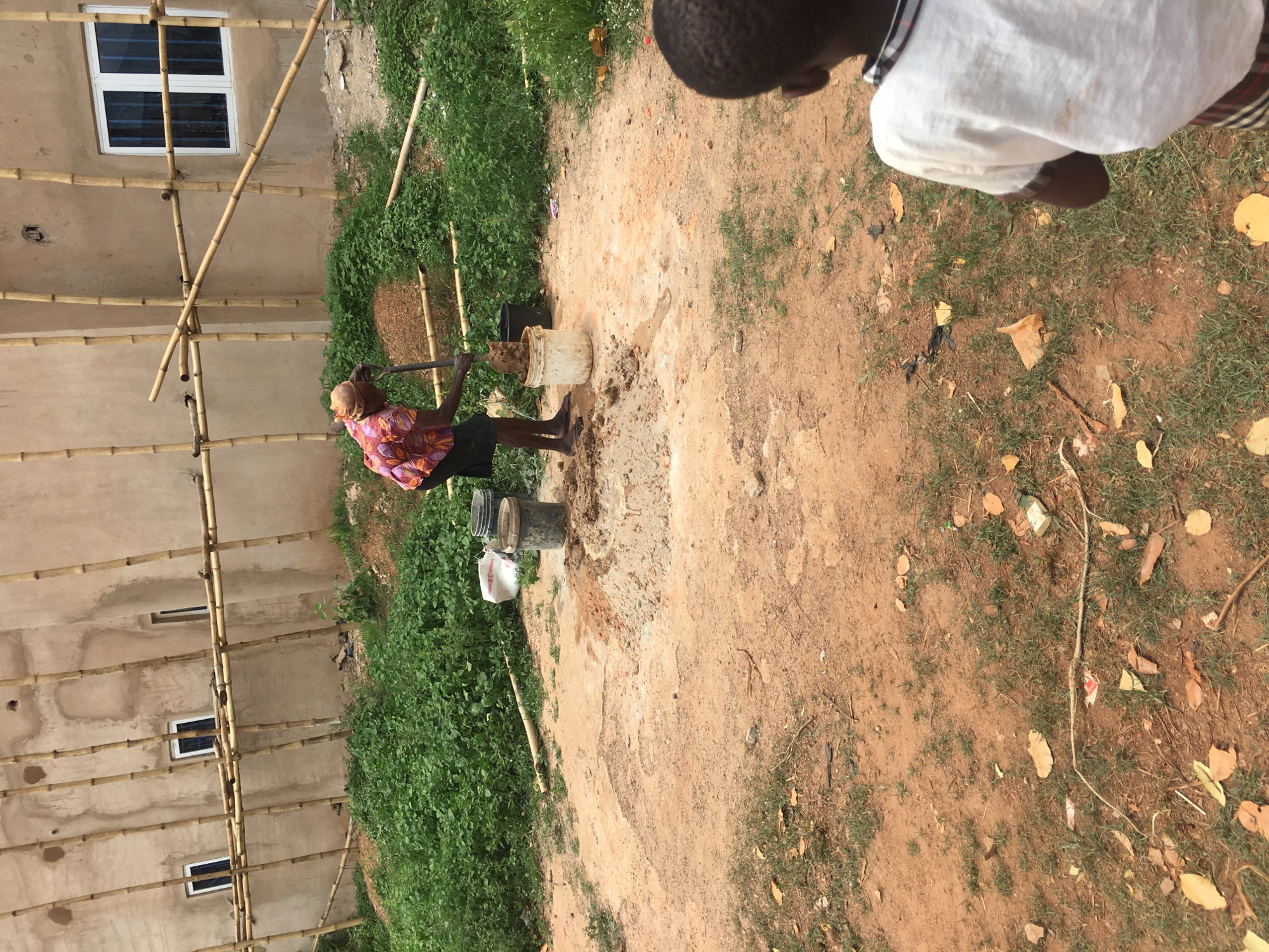 An outdoor scene in Isieke, Nigeria, shows a construction or labor site under natural daylight. In the center, a person with dark skin, wearing a colorful patterned short-sleeved shirt, dark shorts, and a head covering, stands barefoot on reddish-brown dirt. They are holding a long stick or tool, actively mixing what appears to be mud or concrete in a white plastic bucket. Another black bucket is visible nearby.
To the right, in the foreground, the back of a child's head and shoulder are visible, wearing a light-colored top, looking away from the camera.
In the background, to the left, is a light-colored building under construction or renovation. Numerous bamboo poles form scaffolding against its walls, a common construction method in the region. Several white-framed windows with dark panes are visible on the building.
The ground is a mix of reddish-brown earth and patches of green grass or weeds, with some scattered dry leaves and small pieces of litter. The bright lighting suggests it is daytime. No visible text is discernible in the image. The overall scene depicts manual labor associated with construction in a rural or developing setting.