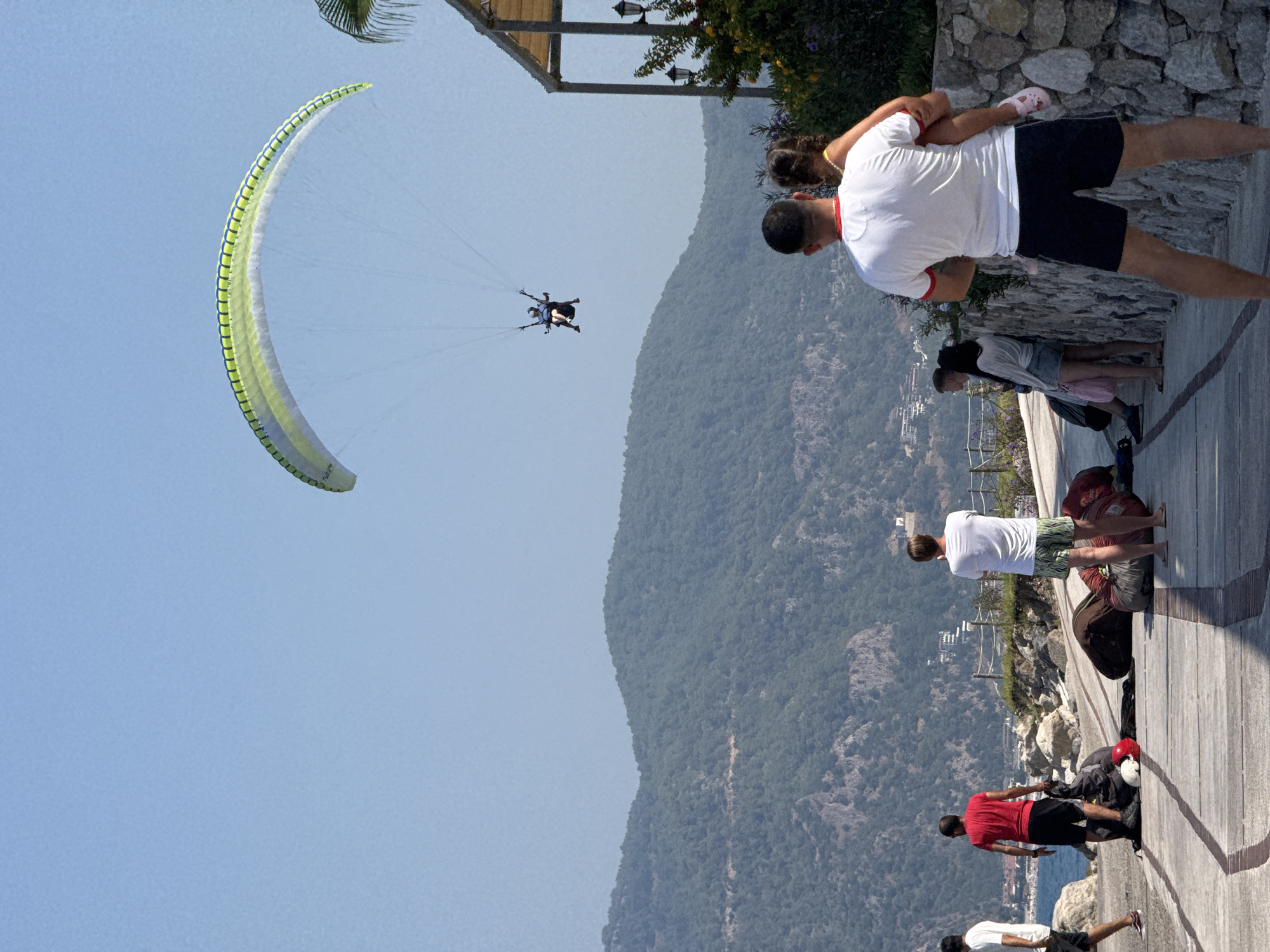 In Fethiye, Turkey, a paraglider is visible in the sky, soaring above a lush, green mountainous landscape under a clear blue sky. Below, on a wooden boardwalk, several people are gathered. A man in a red t-shirt and black shorts stands near large bags, likely paragliding equipment. Another man in a white t-shirt and patterned shorts faces away from the camera. In the foreground, a man wearing a white t-shirt with red trim and dark shorts carries a young girl with her arms around his neck. To the left, another man in a white t-shirt and dark shorts walks away from the camera. The scene suggests a popular paragliding launch or landing area, common in Fethiye, known for its adventure sports.