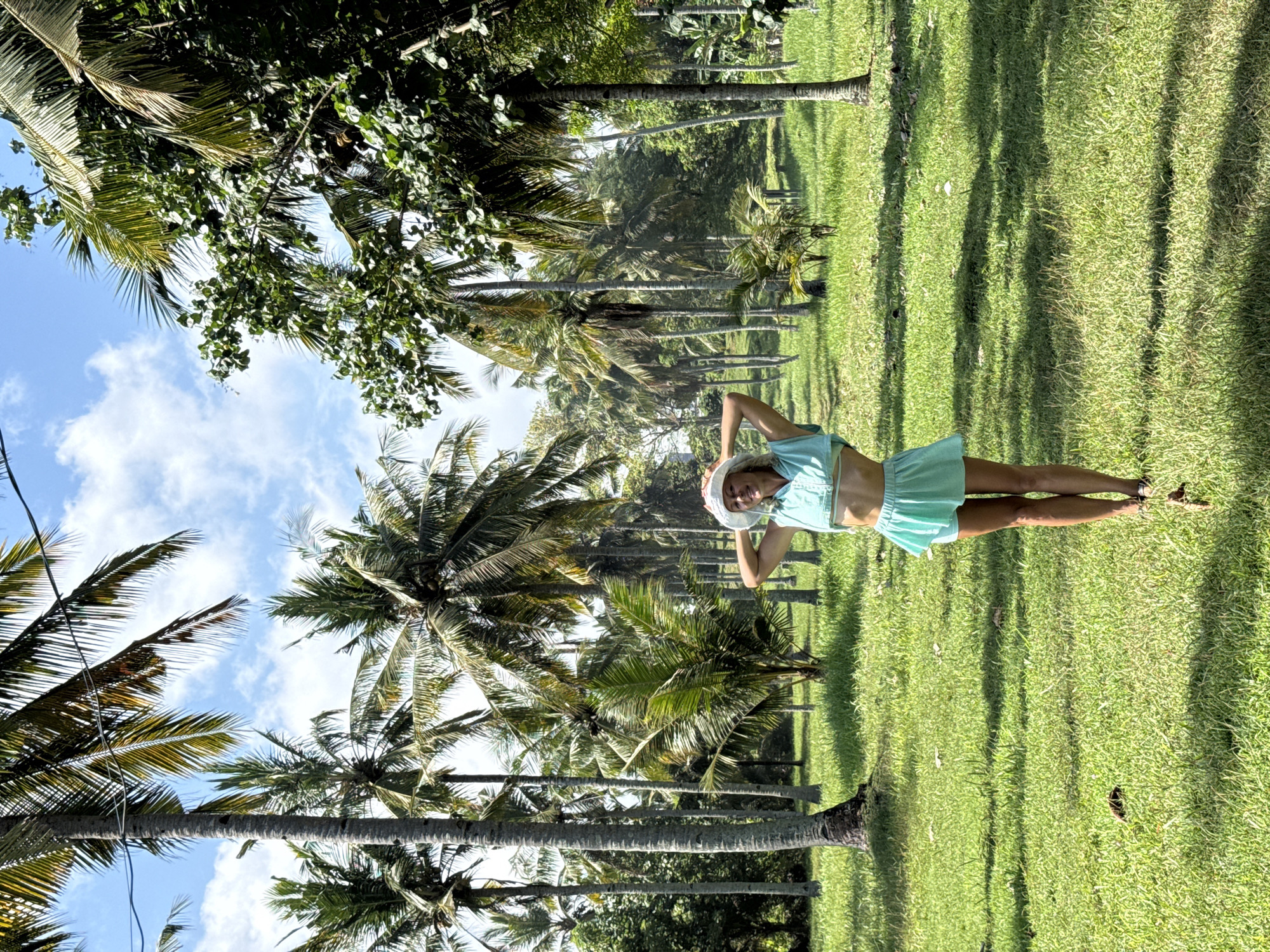 A woman wearing a pale green outfit and white hat is lying on the grass in a location surrounded by palm trees. The sky is visible through the trees and has a few clouds. It is daytime. The location is in the city of Unknown, Unknown.