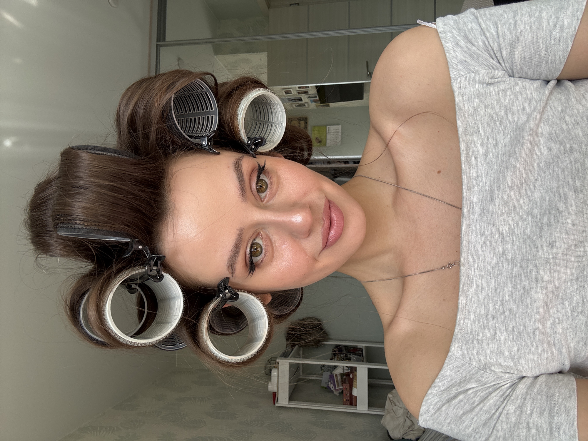 A woman poses indoors, wearing hair rollers. The hair rollers are white and gray. She has brown hair and is wearing a light gray shirt. She is wearing a silver necklace. The location is in Russia. Behind the woman, a glass sliding door, a white shelf, and patterned wallpaper are visible.