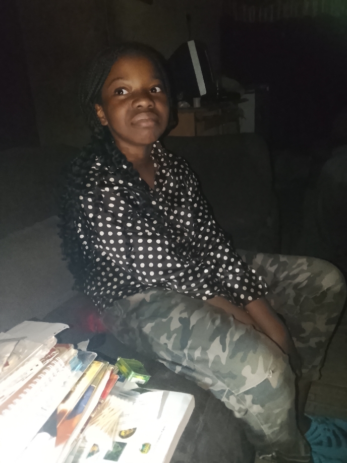 A young girl with braided hair sits on a couch. She wears a black shirt with white polka dots and camouflage pants. In front of her, a stack of books rests on a white surface, and there's a small, green rectangular object nearby. An old television sits in the background. The room is dimly lit. The girl appears to be looking slightly to the side.