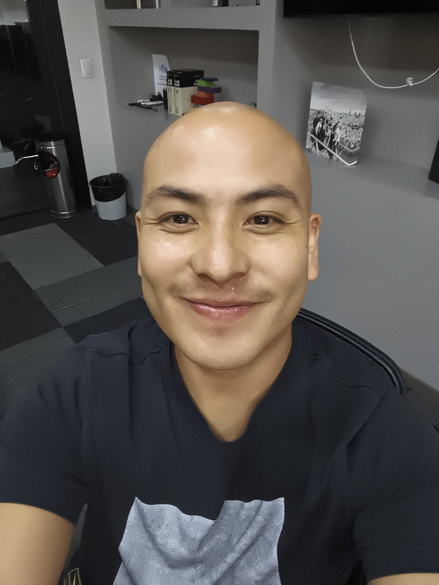 A bald man with brown eyes and a nose piercing is smiling at the camera. He is wearing a dark blue t-shirt with a gray patch on the front. The background is a gray wall with a shelf containing various items, including a black and white photograph. The floor is covered in a gray and black patterned carpet. There is a stainless steel trash can and a black trash can in the background. The man appears to be indoors, possibly in an office setting. There is no information in the image to determine if the location is Mexico City, Mexico.