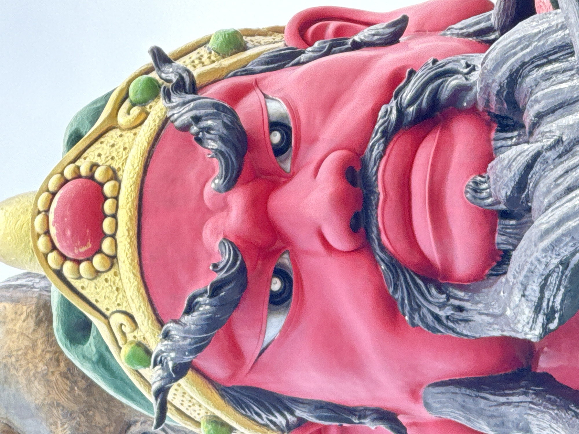 The image is a close-up, frontal view of a statue's face, likely a deity or guardian figure, characterized by its vibrant red skin, large, expressive eyes with dark irises and white pupils, and a prominent black mustache and beard. The figure wears an ornate golden headdress adorned with red and yellow circular elements, and a green cap-like structure. Black, stylized hair or decorative elements frame the face, extending from the temples and chin. The statue appears to be located in Baan Hua Thanon, Thailand. The background is a bright, uniform sky.