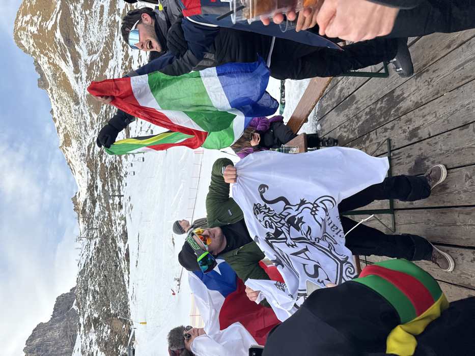 The image shows a vibrant outdoor gathering of people on a wooden deck at a ski resort, set against a backdrop of snow-dusted mountains and a ski slope in Alagna Valsesia, Italy. The weather appears clear and sunny.
In the upper part of the frame, a man wearing ski goggles on his head and a dark jacket is laughing heartily, holding a flag resembling the South African flag with its distinctive red, green, white, blue, and yellow colors. His hand, alongside others, holds plastic cups filled with beverages, indicating a festive toast or celebration. Below him, another man in a green jacket and ski goggles holds a large white flag emblazoned with a black crest featuring a rampant lion and partially visible text that reads "Valsesian". Another flag, red, white, and blue, is also visible near him.
Several other individuals are part of the group, dressed in winter clothing suitable for the mountain environment. Some are seated on a wooden bench, while others stand and socialize. Notable accessories include a fur-trimmed hat and a colorful red, green, and yellow striped beanie. A ski lift structure is discernible in the distant background, reinforcing the setting. The overall mood is jovial and celebratory, with people enjoying drinks and each other's company amidst the stunning alpine scenery.
