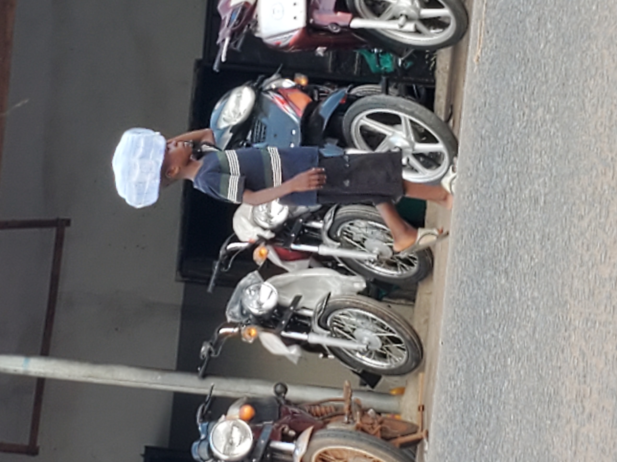This image, taken in Mararaba, Nigeria, captures a daytime scene featuring a young boy standing alongside a row of parked motorcycles.
The central subject is the boy, who appears to be an adolescent. He is dressed in a blue polo shirt with horizontal white and dark green stripes, dark shorts, and light-colored sandals. A distinct feature is the light-colored, possibly white or light blue, net-like cap or object resting on his head. He stands in a casual posture, looking towards the right side of the frame with a neutral and observant expression.
To his left, several motorcycles are neatly lined up, suggesting they are either for sale, awaiting repair, or simply parked in a common area. At least five motorcycles are clearly visible, displaying a variety of colors including red, blue, white, and a darker, possibly maroon or brown tone. They exhibit different designs, with some appearing newer and well-maintained, featuring chrome accents and modern alloy wheels, while others show signs of age or wear, like the one with spoked wheels at the bottom.
The setting appears to be an outdoor urban or semi-urban environment. A textured grey surface, consistent with asphalt or concrete pavement, is visible on the right side of the image, suggesting a street or pathway. On the far left, there's a glimpse of a light-colored wall or building structure with dark vertical elements, possibly pillars or frames. The bright, even lighting indicates it is daytime, likely under clear skies. There is no visible text in the image.